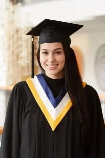 Young woman with long dark hair smiling outdoors beside a tree and green leafy bushes, wearing sunglasses on her head and a black and white checkered tank top.
