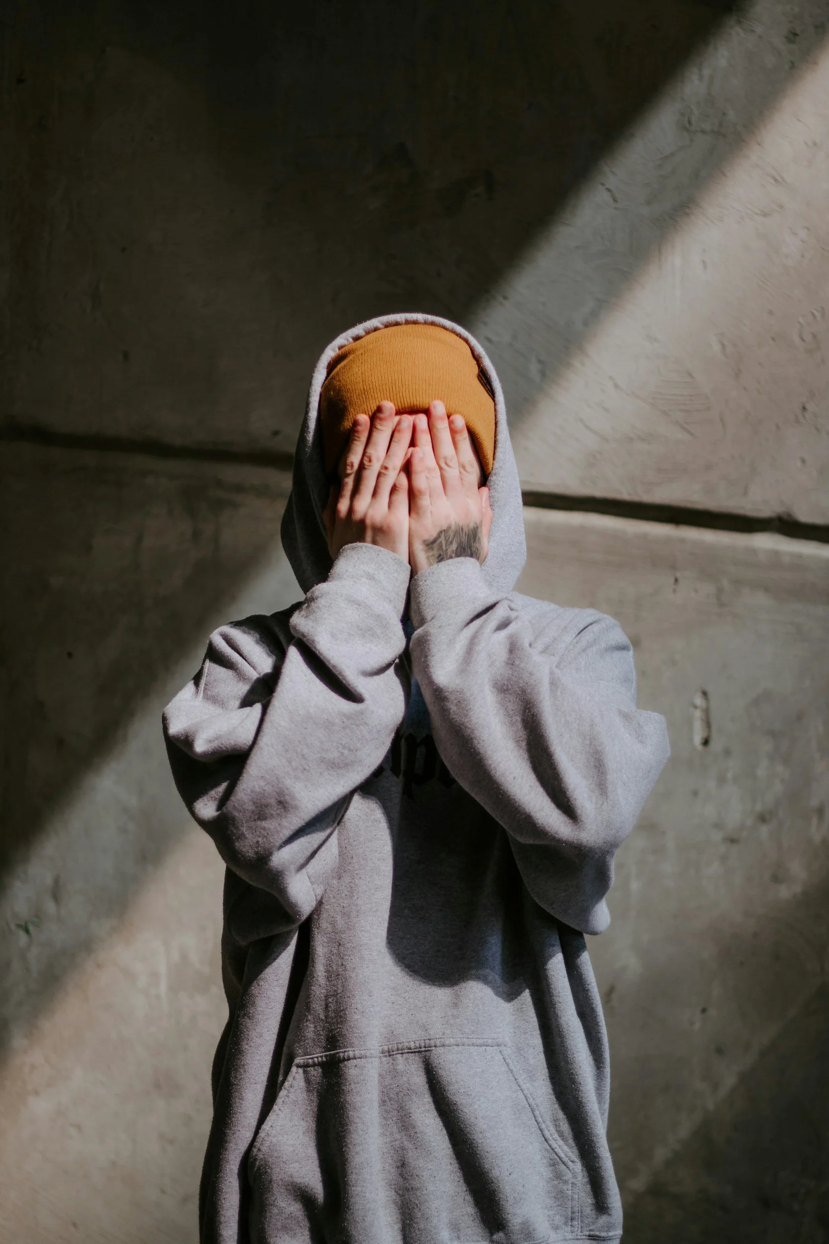 Person wearing a gray hoodie and a brown beanie covering their face, standing against a concrete wall with a diagonal shadow.