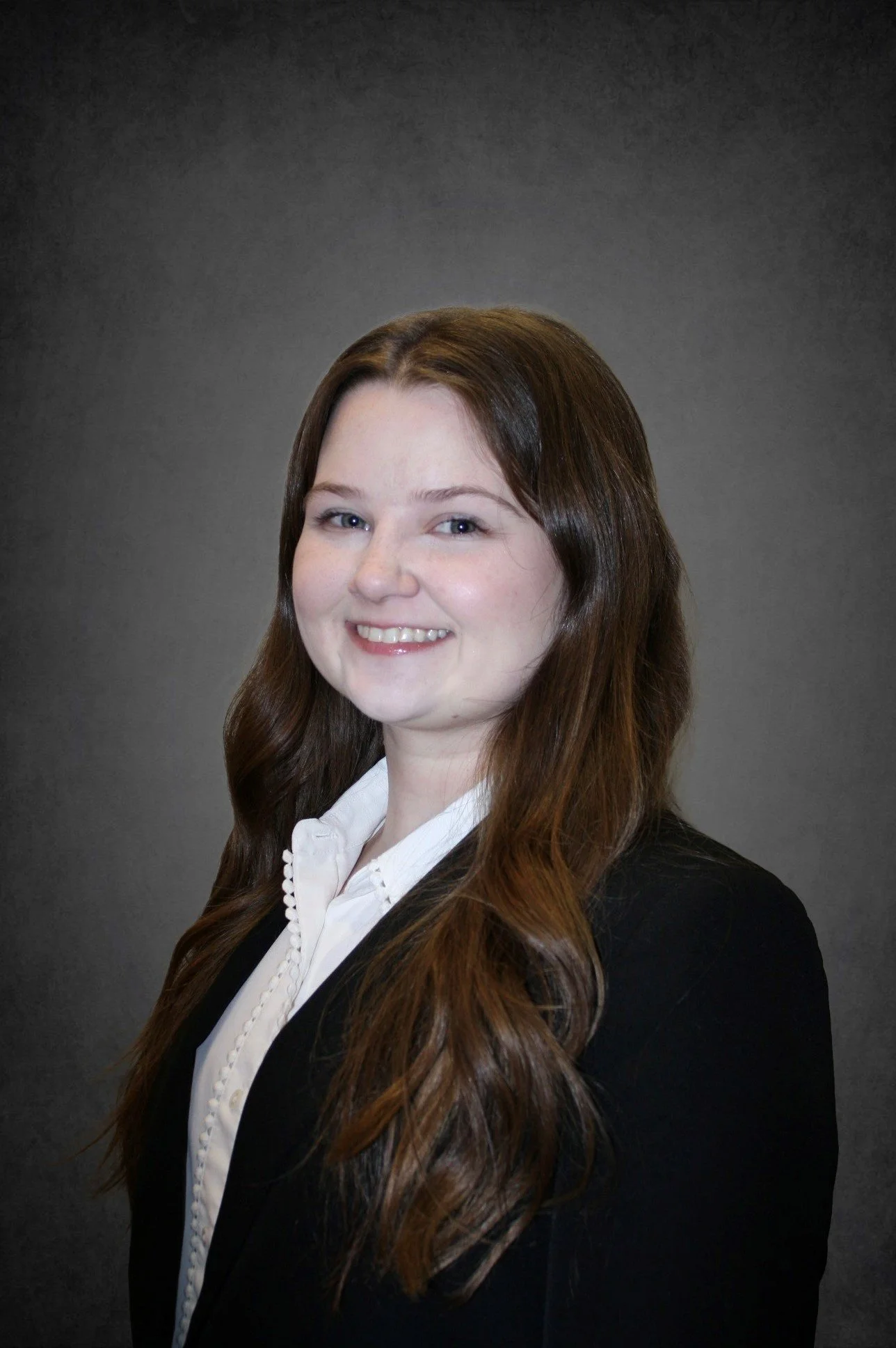 A professional portrait of a young woman with long brown hair, wearing a black blazer and white blouse, smiling against a gray background.