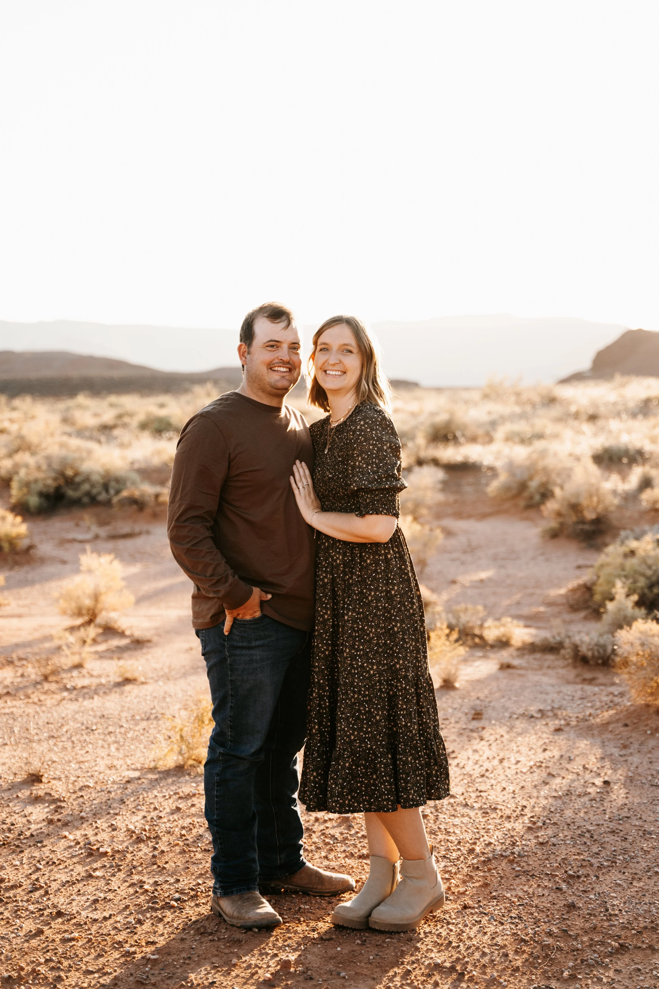 A couple standing close together in a desert landscape during sunset, smiling and looking at the camera, with mountains in the background.