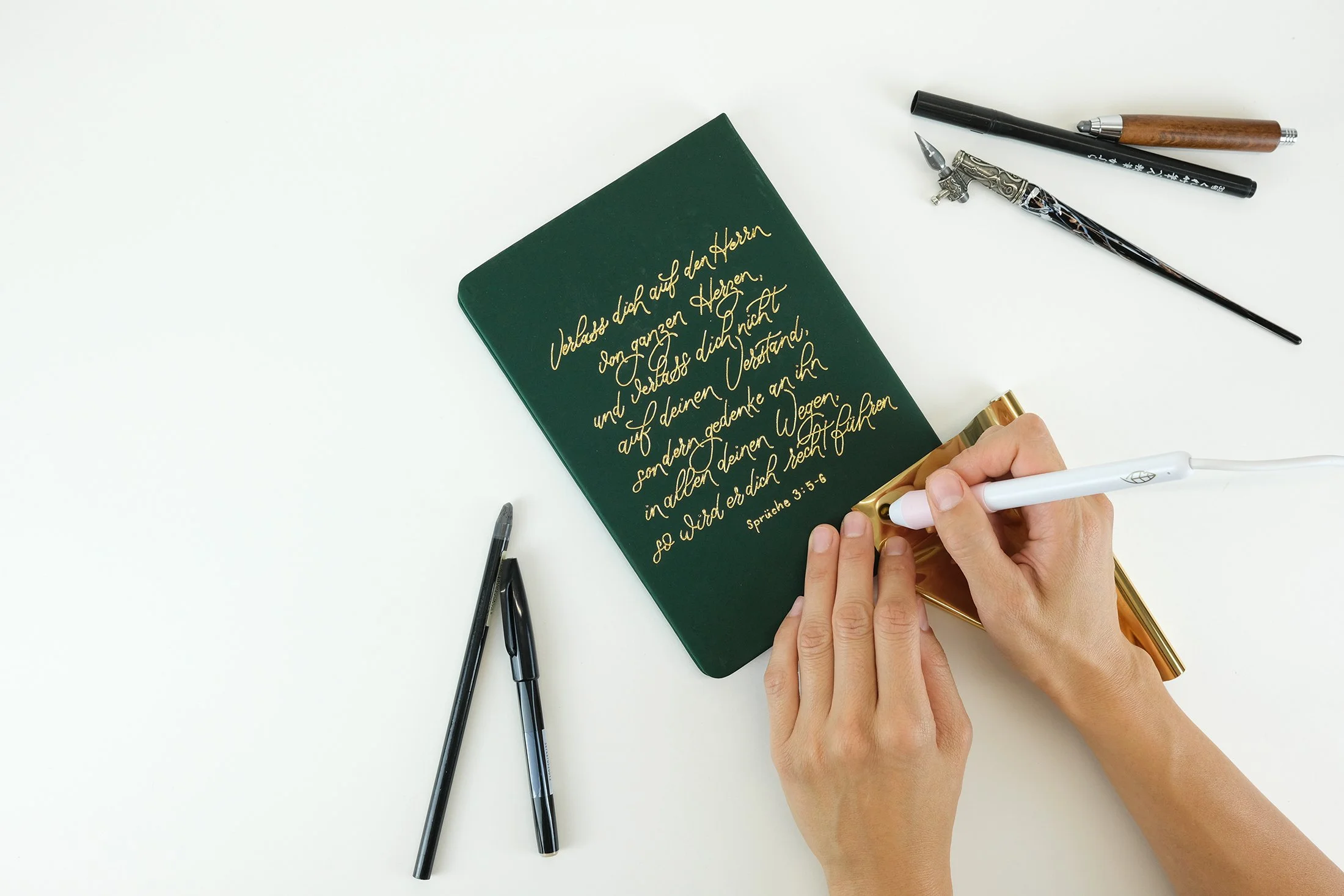 Person schreibt mit einem Kalligraphiestift auf grünes Notizbuch, umgeben von Stiften und Kalligraphie-Werkzeugen auf weißem Tisch.