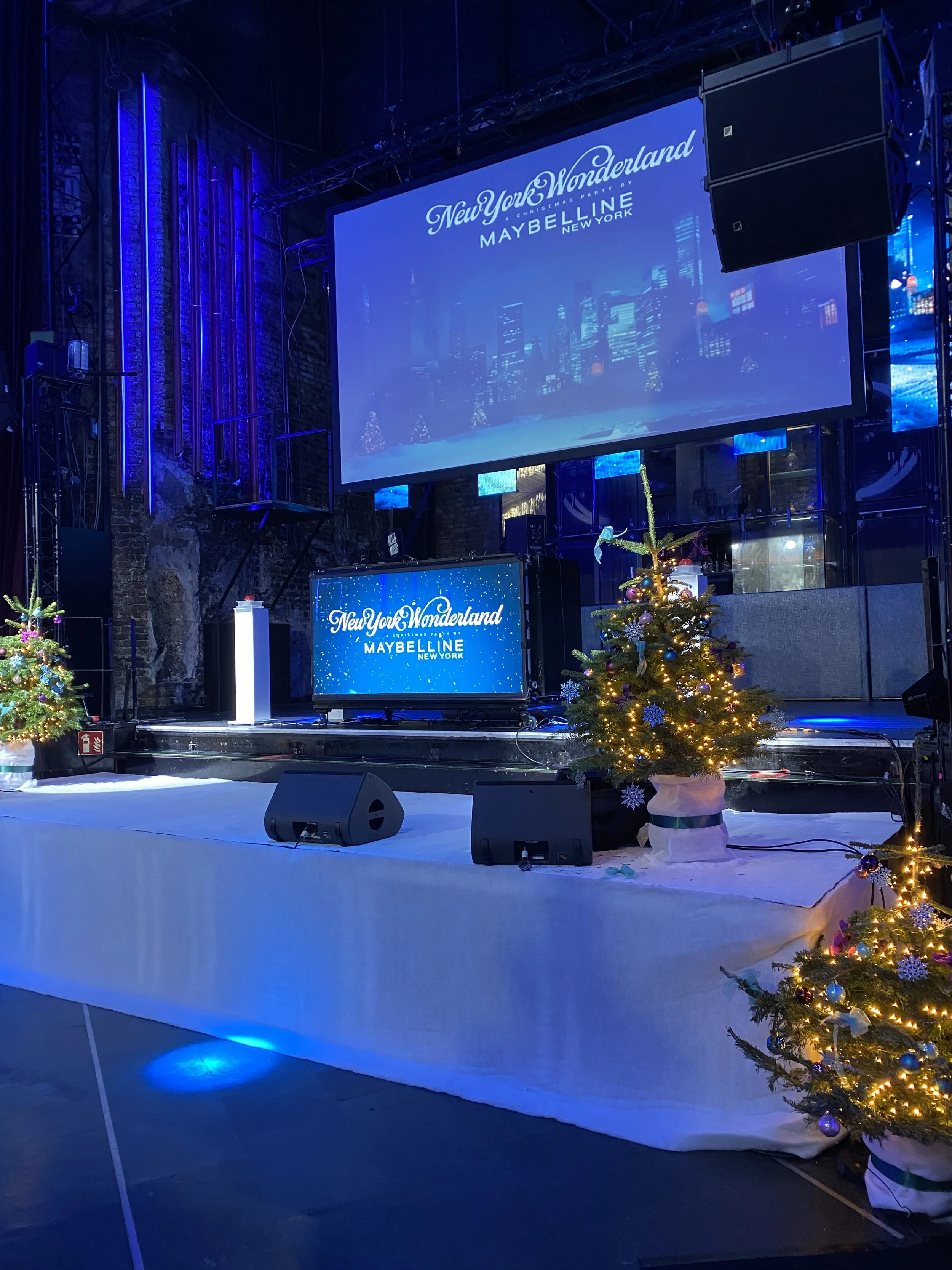 Bühne mit New York Wonderland- und Maybelline-Werbebanner, Dekoration mit Weihnachtsbäumen und Lichtern, Lautsprecher und Mikrofone am Boden.