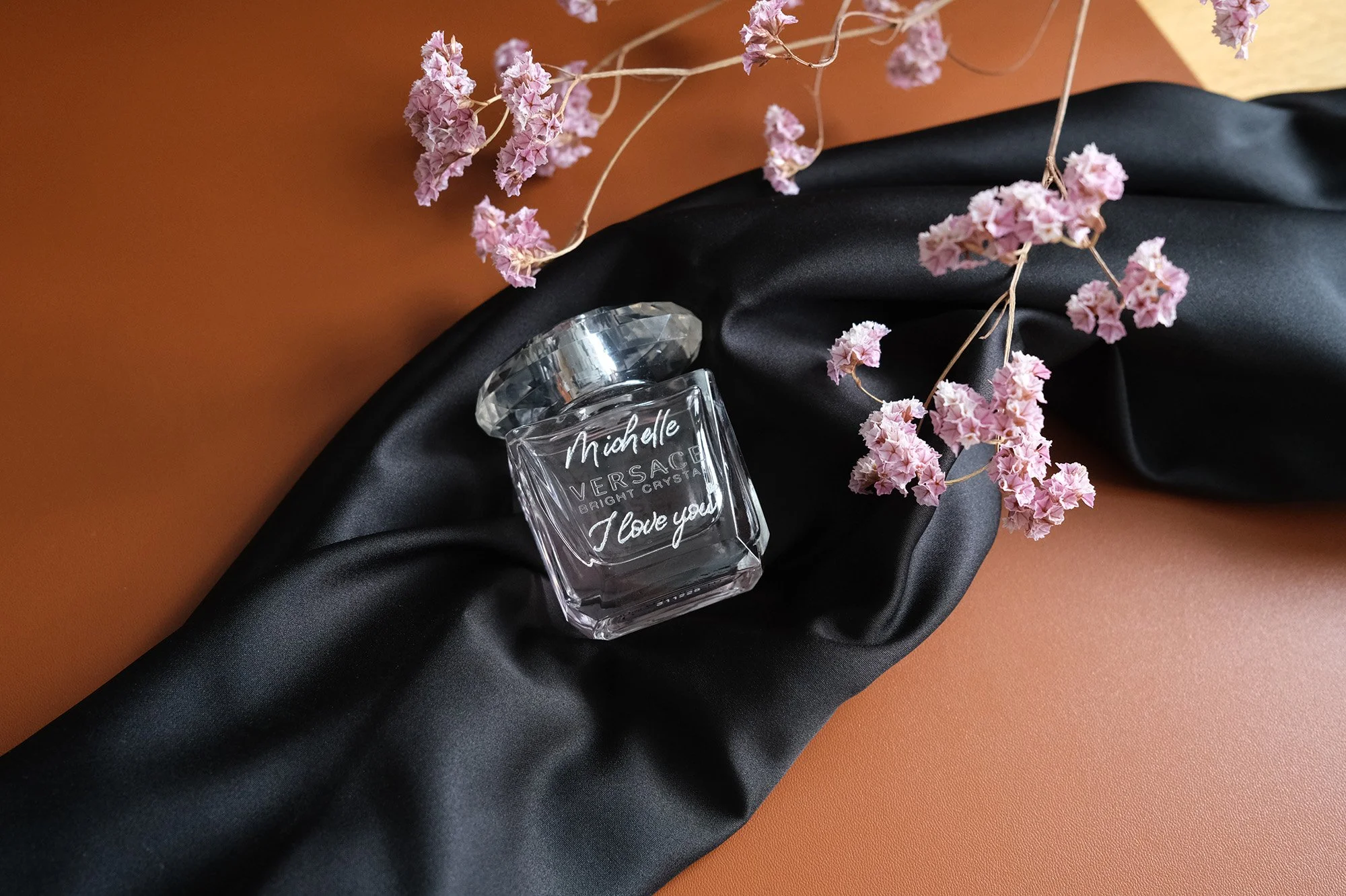 Parfümflasche Versace Bright Crystal auf schwarzem Stoff mit rosa Trockenblumen.