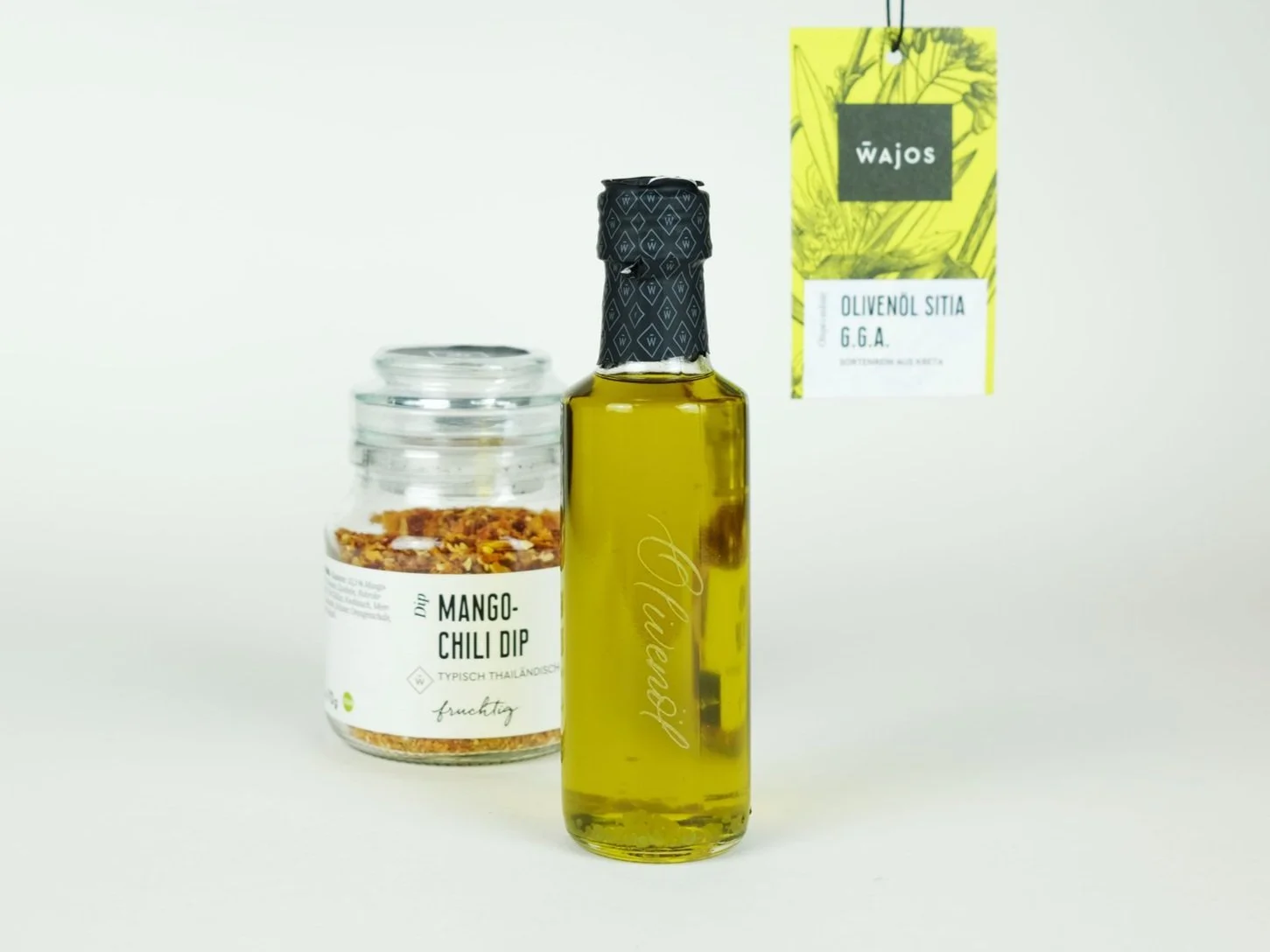 Eine Flasche Olivenöl von Wajos steht neben einem Glas Mango-Chili-Dip. Das Etikett auf der Flasche zeigt Olivenöl Sitia G.G.A. Das Glas hat ein Etikett mit der Aufschrift Mango-Chili Dip, typisch thailändisch, fruchtig.