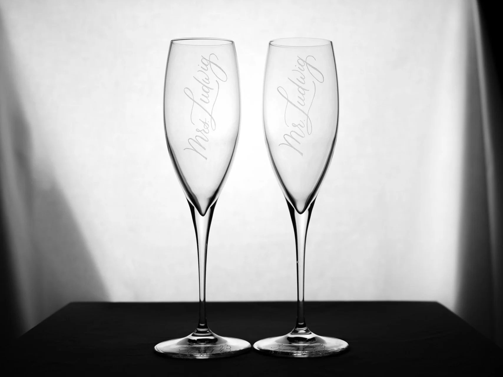 Zwei leere Sektgläser mit Gravur "Mr. Ludwig" und "Mrs. Ludwig" auf einem Tisch vor einem neutralen Hintergrund.