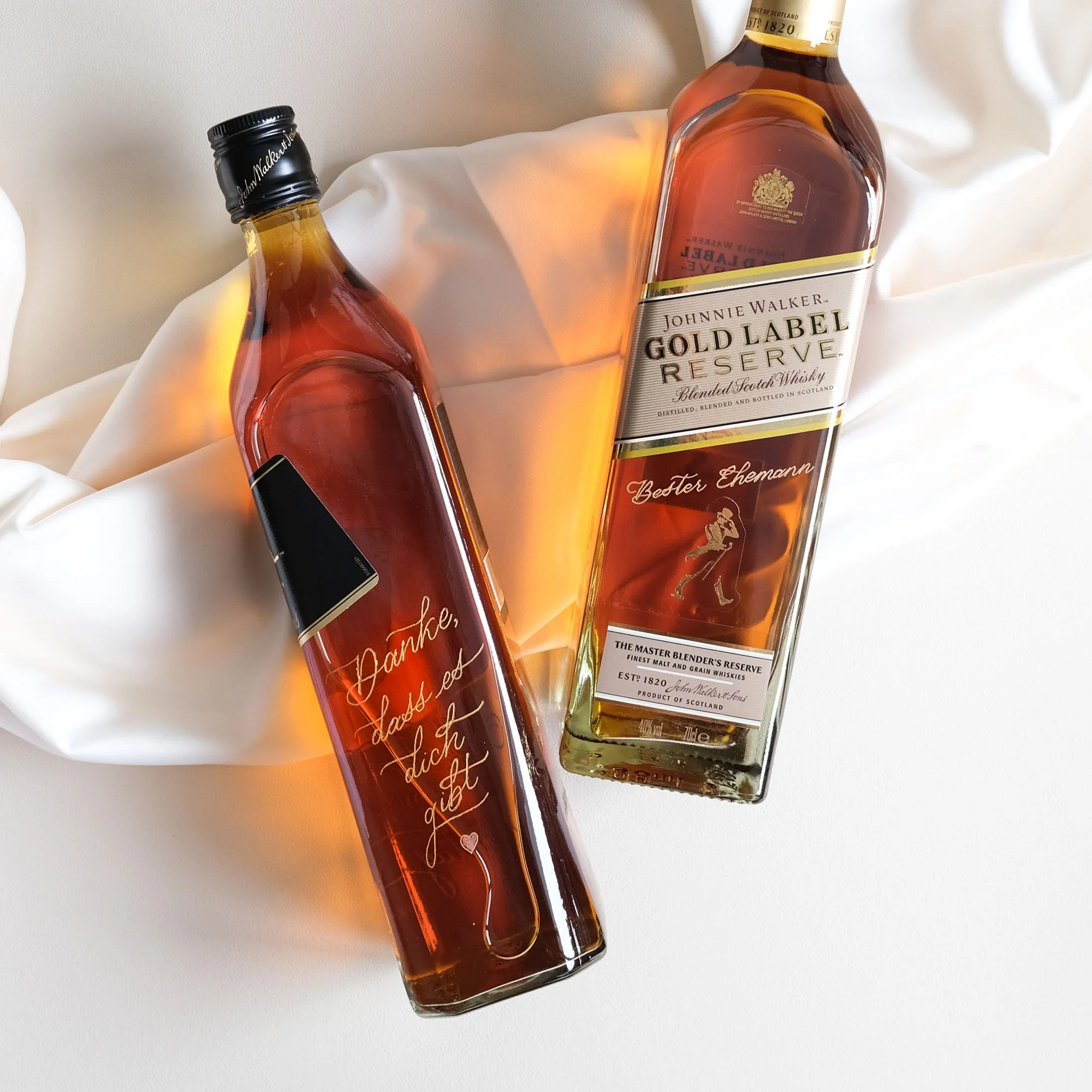 Zwei Whiskyflaschen auf einem weißen Stoff. Die linke Flasche hat eine persönliche Botschaft in deutscher Sprache, die rechte Flasche ist ein Johnnie Walker Gold Label Reserve.