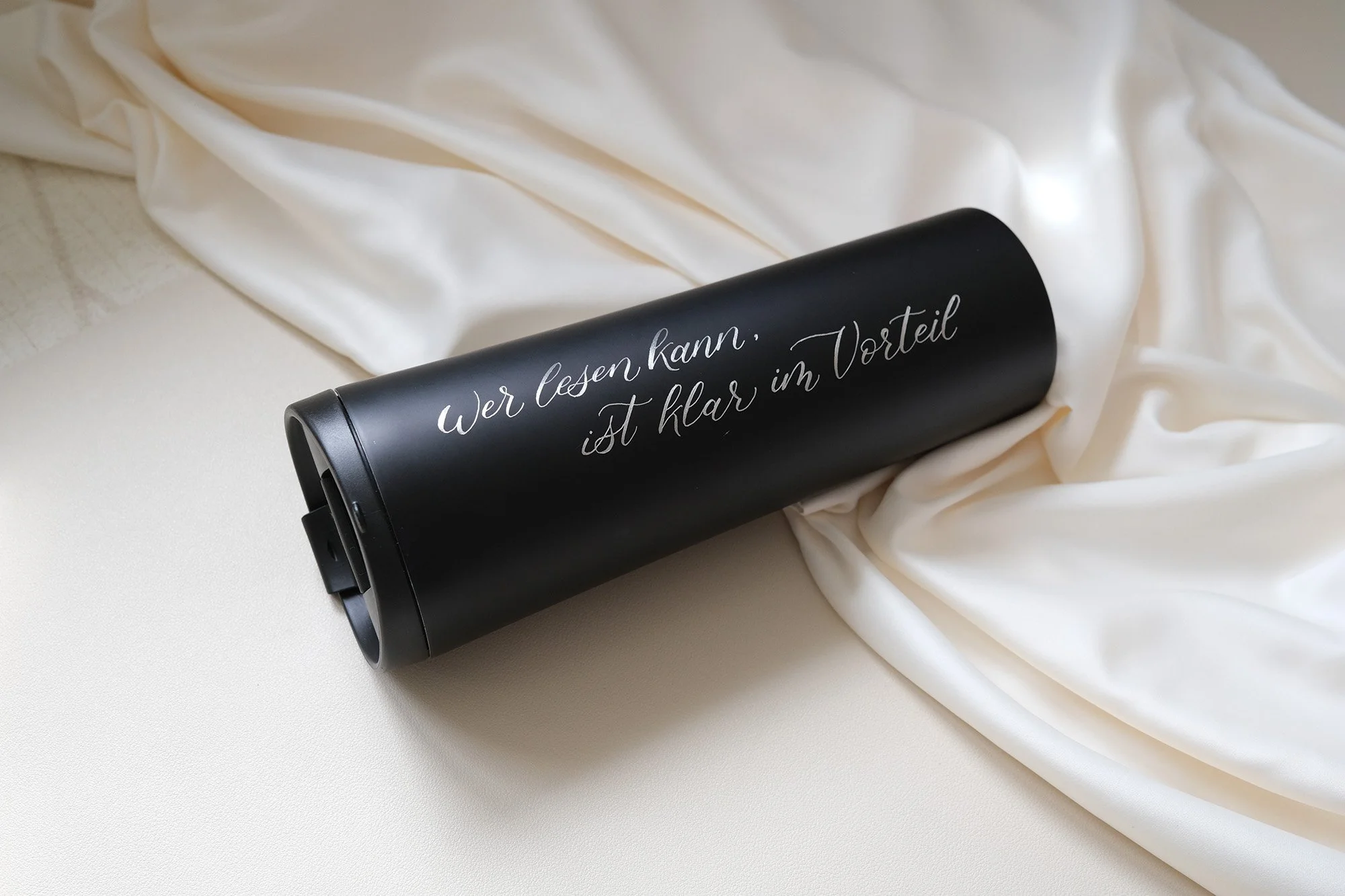 Schwarze Thermosflasche mit Aufschrift "Wer lesen kann, ist klar im Vorteil" auf weißem Stoff.