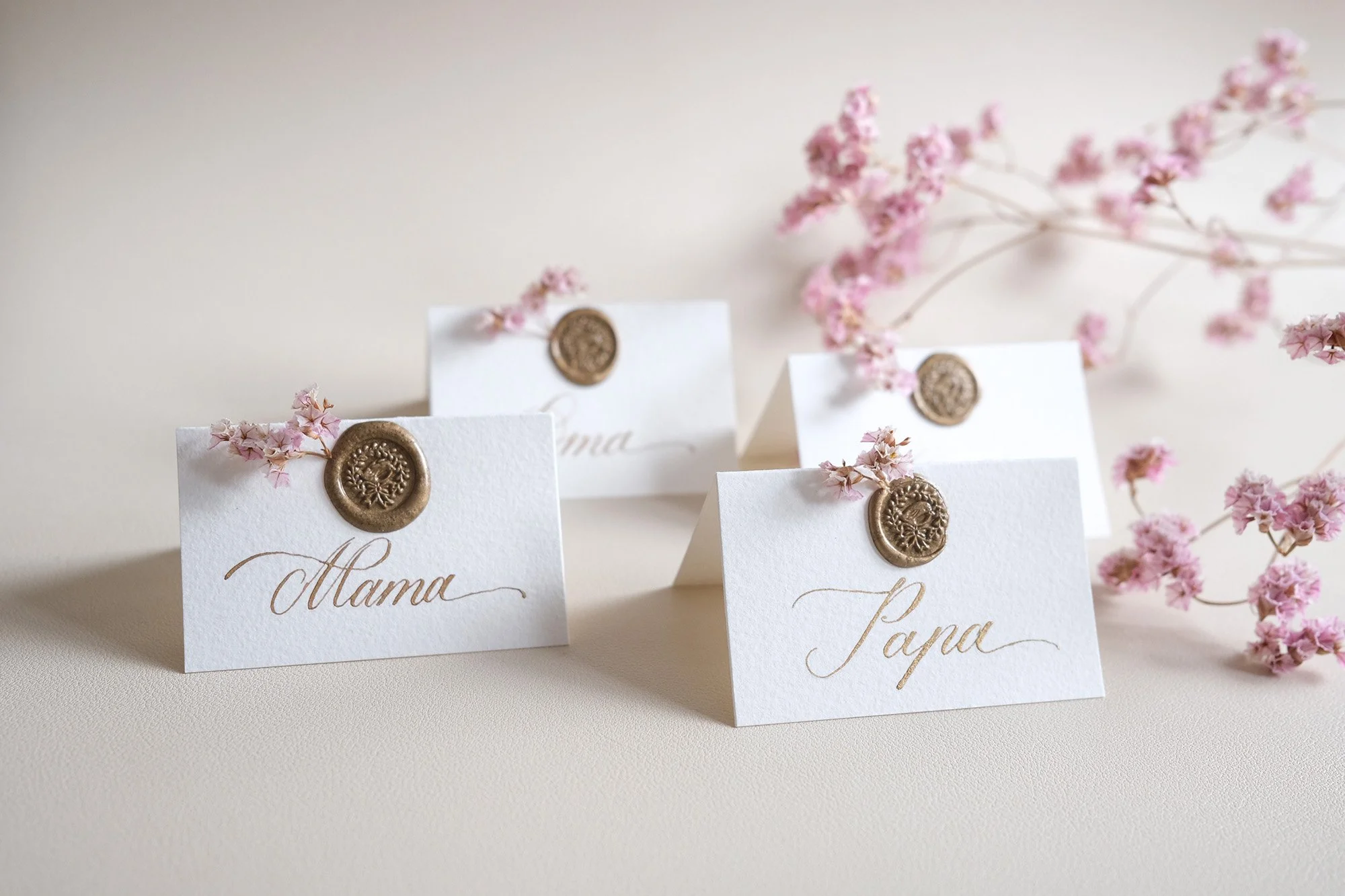 Handgeschriebene Tischkarten mit "Mama" und "Papa" in Kalligrafie, verziert mit Wachssiegel und kleinen rosa Blüten.