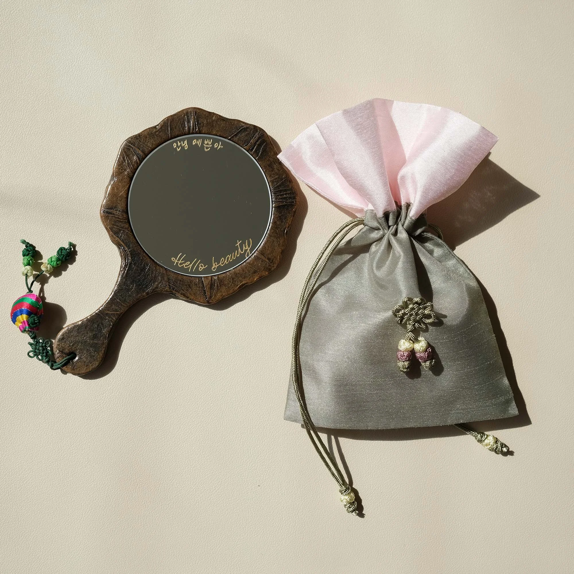 Handspiegel mit Holzrahmen und dekorativem Anhänger neben einem Seidenbeutel mit Knotenverschluss auf beigem Hintergrund.