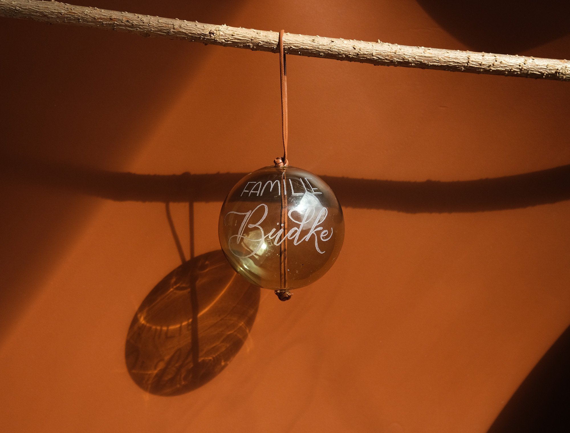 Glaskugel mit Aufschrift "Familie" und einem unscharfen Namen, aufgehängt an einem Ast, mit Schatten auf einer orangen Oberfläche.
