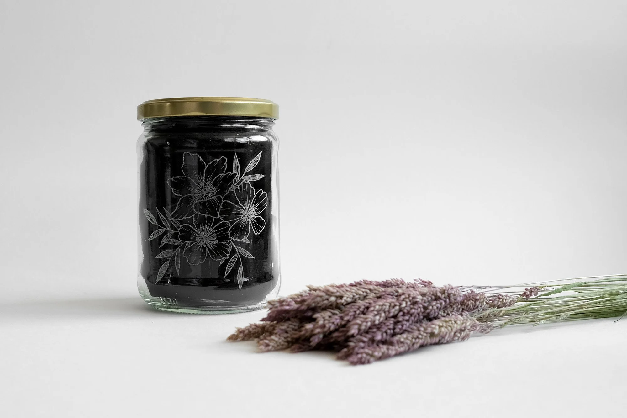 Ein Glas mit schwarzem Inhalt und goldenem Deckel, verziert mit Blumenmuster, neben einem Bündel lila getrocknetem Lavendel auf hellem Hintergrund.