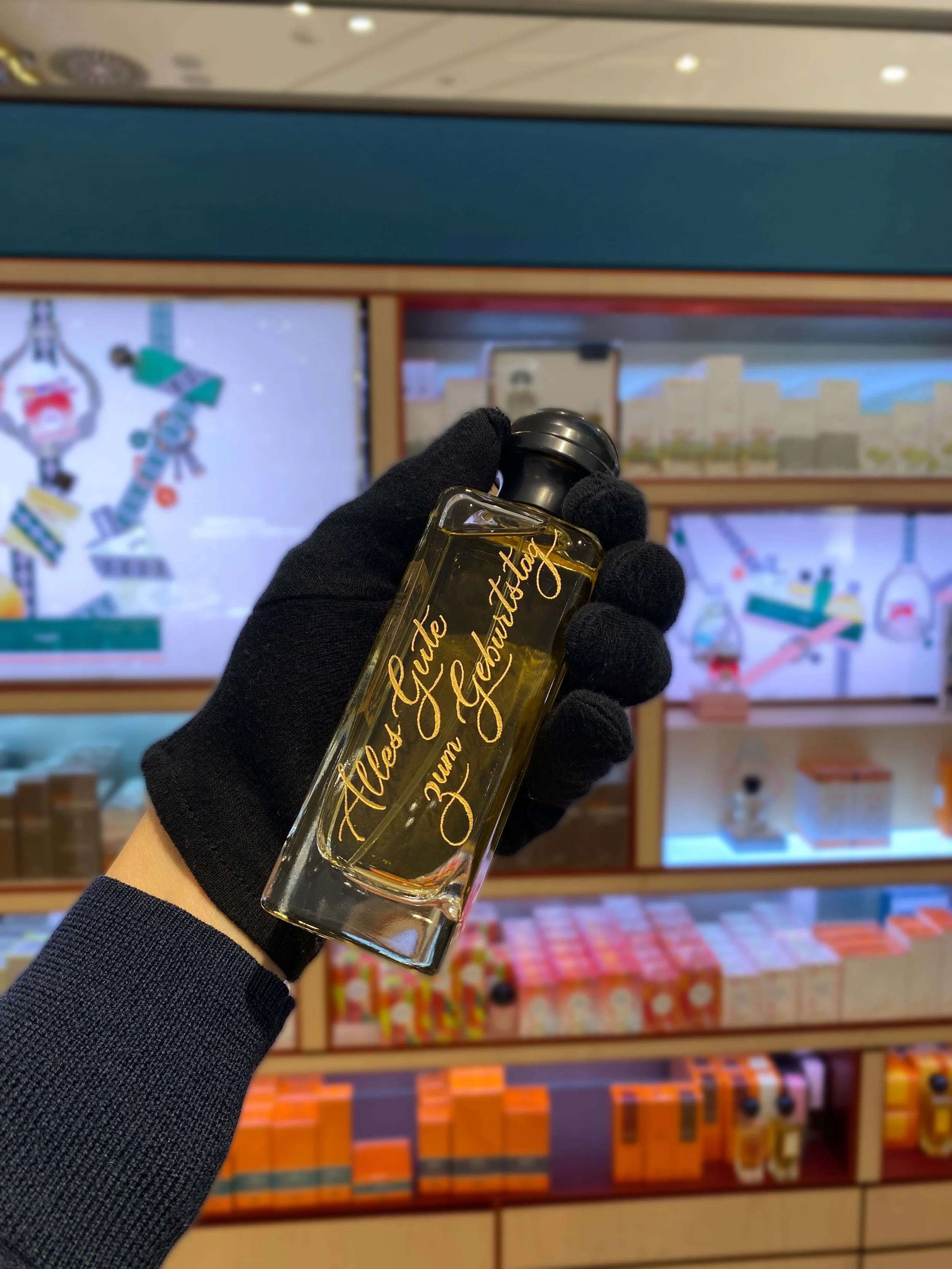 Eine Person hält eine Flasche mit der Aufschrift "Alles Gute zum Geburtstag" vor einem Regal mit Geschenken und Verpackungen.