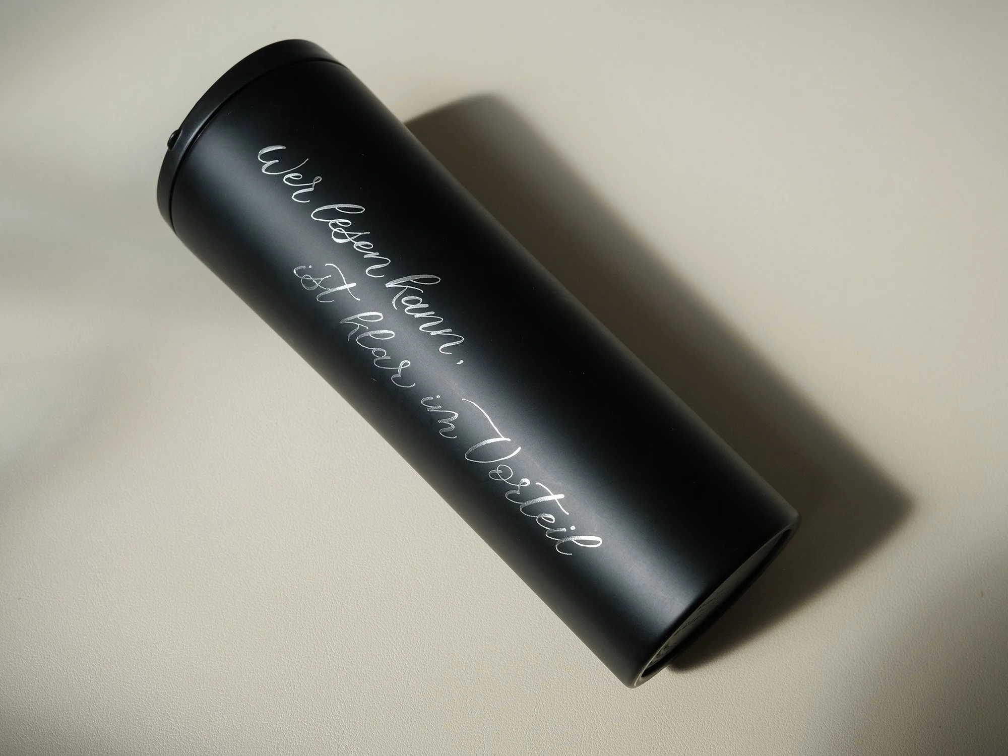 Schwarzer Thermobecher mit der Aufschrift 'Wer lesen kann, ist klar im Vorteil' auf einer hellen Oberfläche liegend.