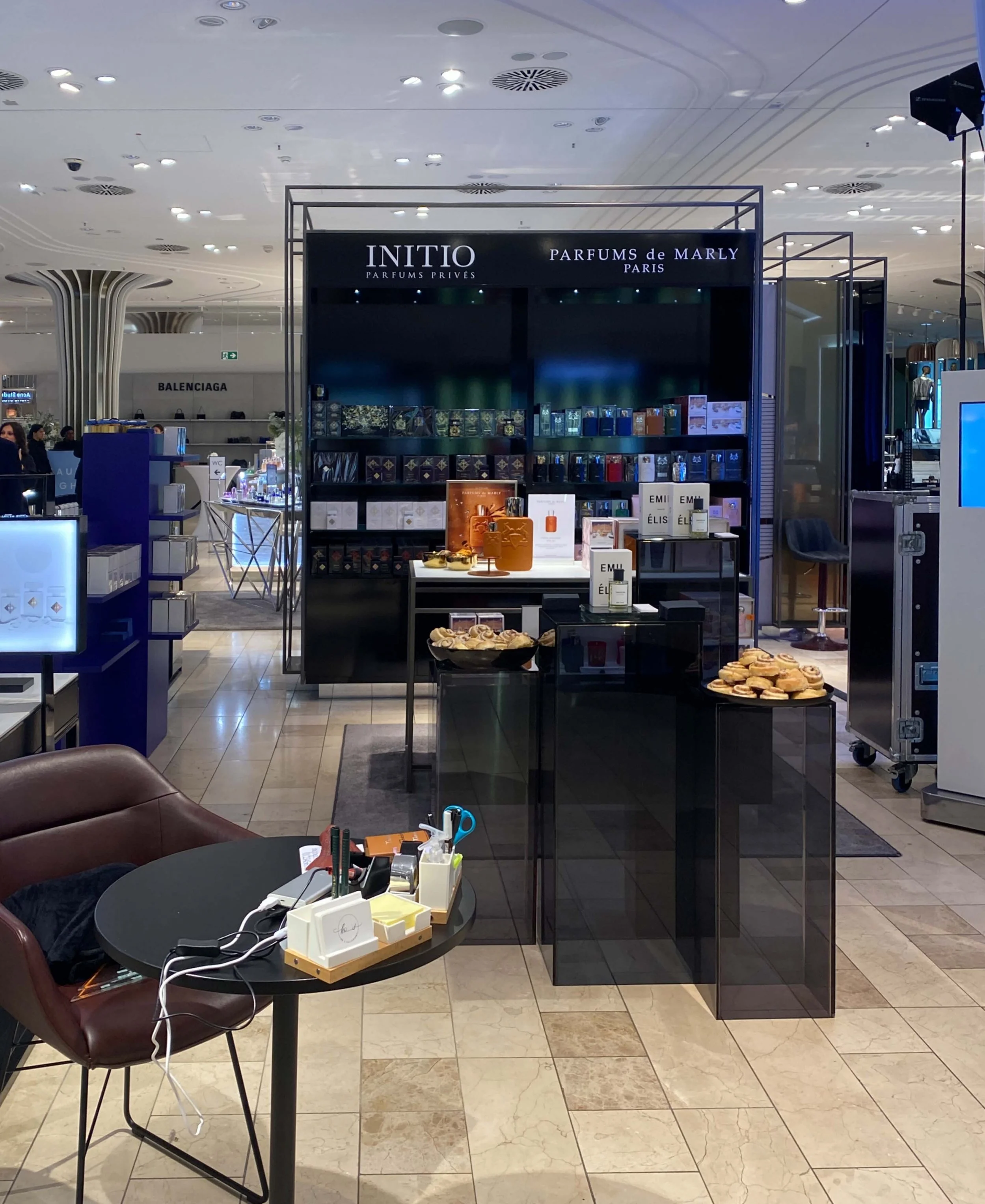 Innenbereich eines Kaufhauses mit einem Parfümstand der Marken Initio und Parfums de Marly, dekoriert mit Parfümflaschen und Snacks. Im Vordergrund steht ein runder Tisch mit Büromaterial.