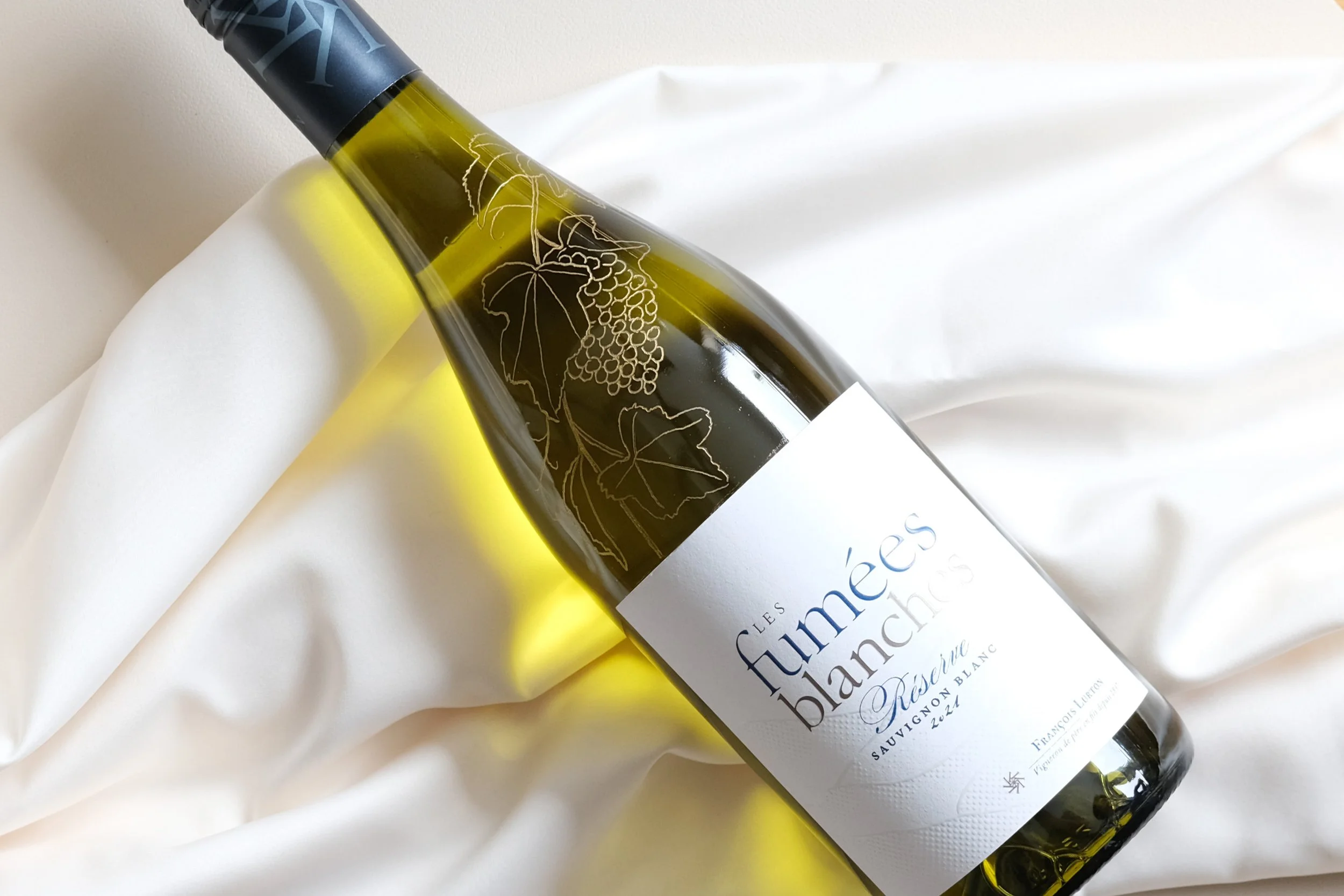 Flasche Weißwein "Les Fumées Blanches" auf weißem Stoff liegend.