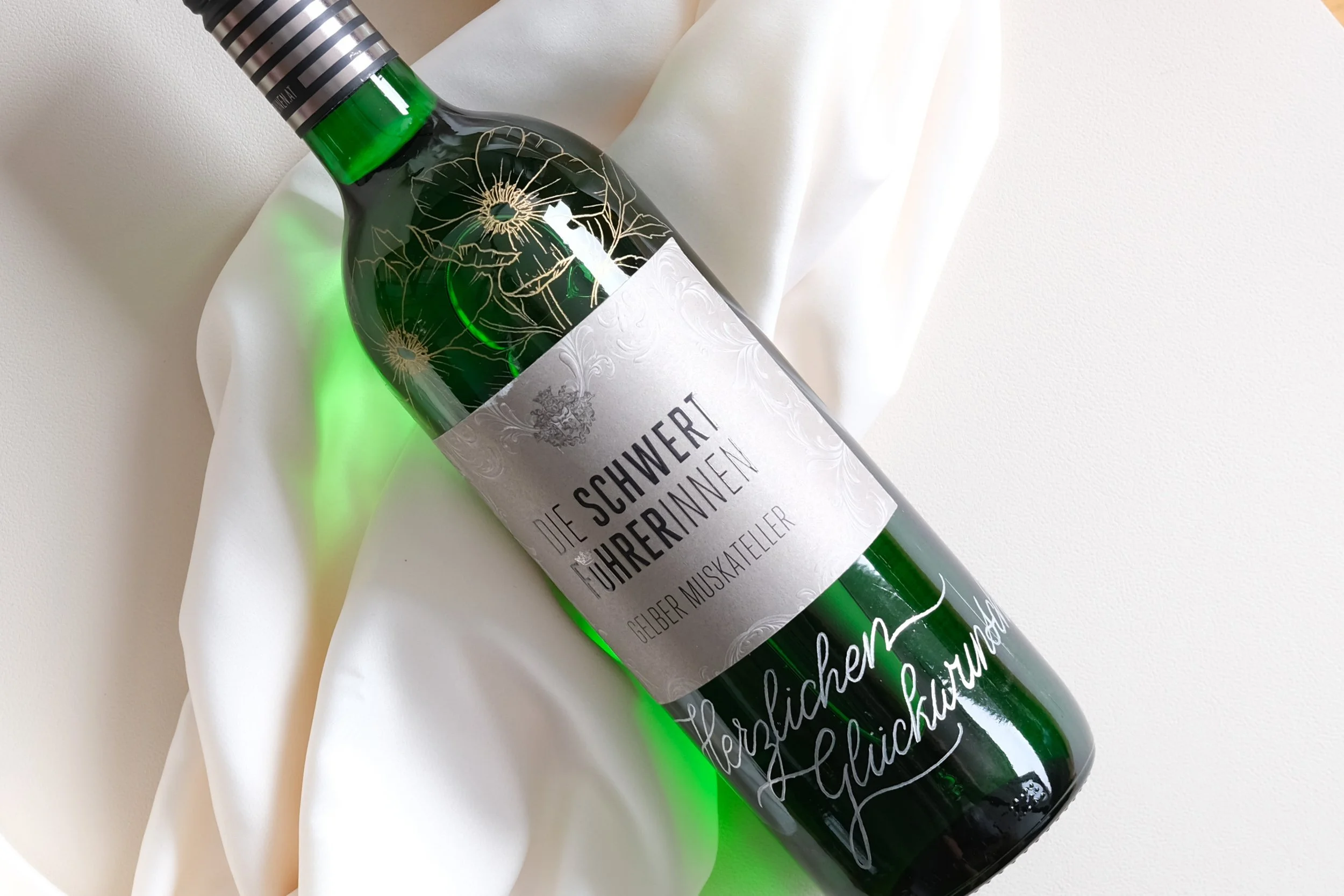 Grüne Weinflasche mit dem Etikett "Die Schwertführerinnen Gelber Muskateller", dekoriert mit Blumenmuster auf hellem Stoff.