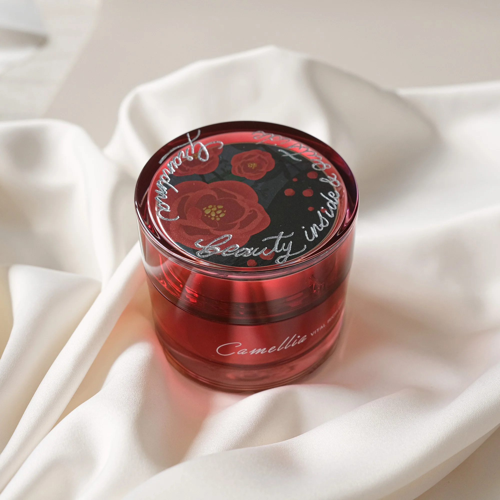 Roter Kosmetiktiegel mit Blütendesign auf dem Deckel, umgeben von weißem Stoff.