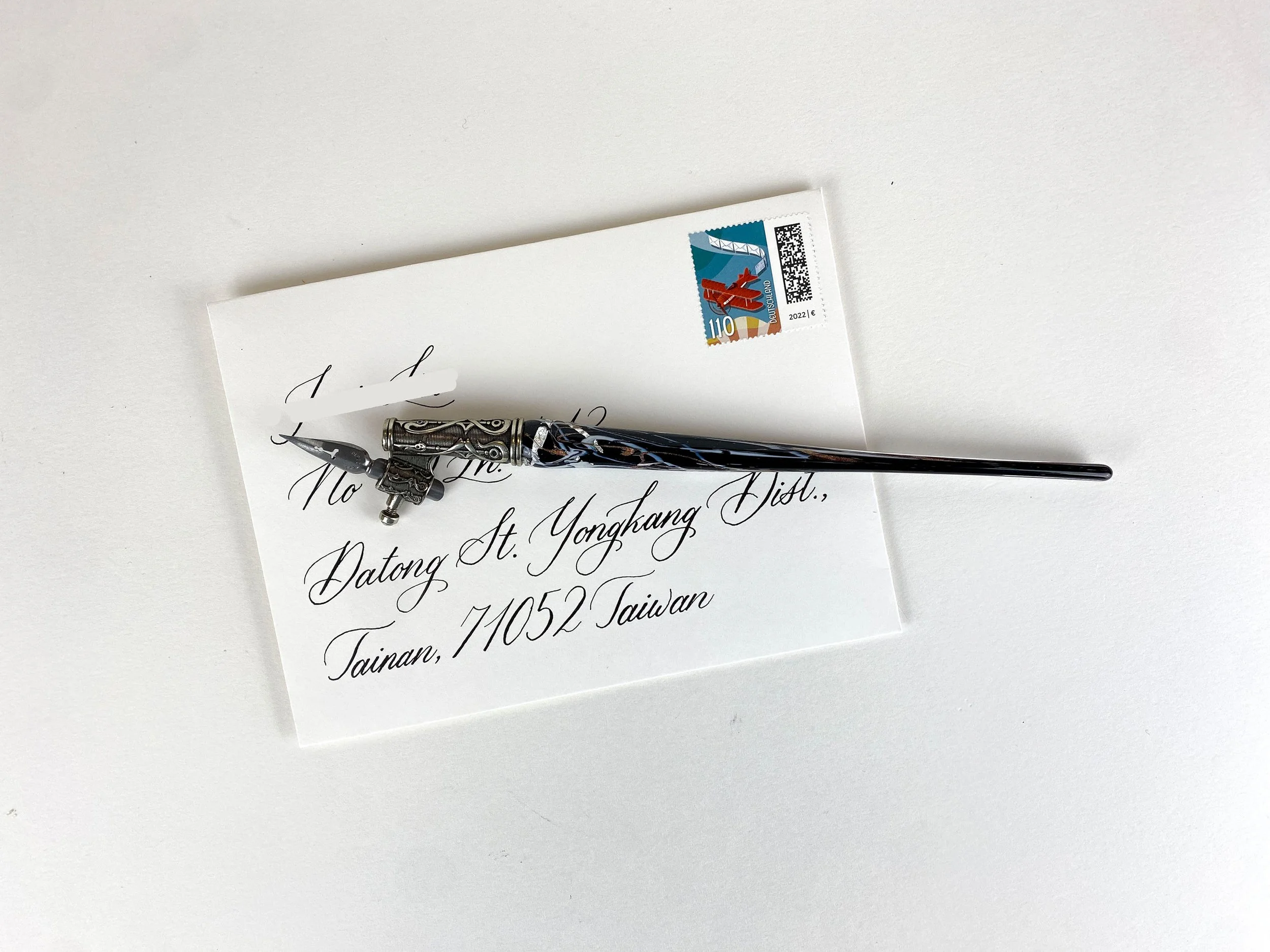 Eine mit Kalligraphie beschriebene weiße Briefhülle mit einer Schreibfeder und einer Briefmarke oben drauf.