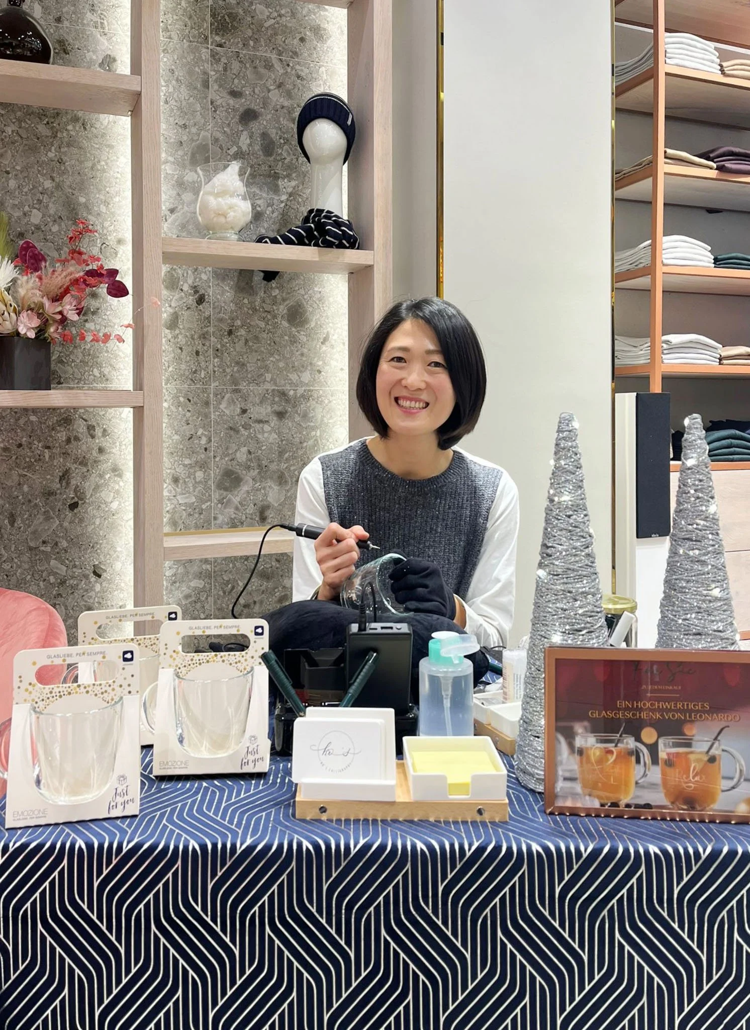 Eine Frau sitzt an einem Tisch in einem Geschäft und graviert ein Glas. Der Tisch ist mit einem dunkelblauen Tuch bedeckt, auf dem sich verschiedene Artikel, darunter Gläser und Dekorationen, befinden. Im Hintergrund sind Regale mit Kleidung und Dekorationen zu sehen. Die Frau lächelt und trägt einen Pullover.
