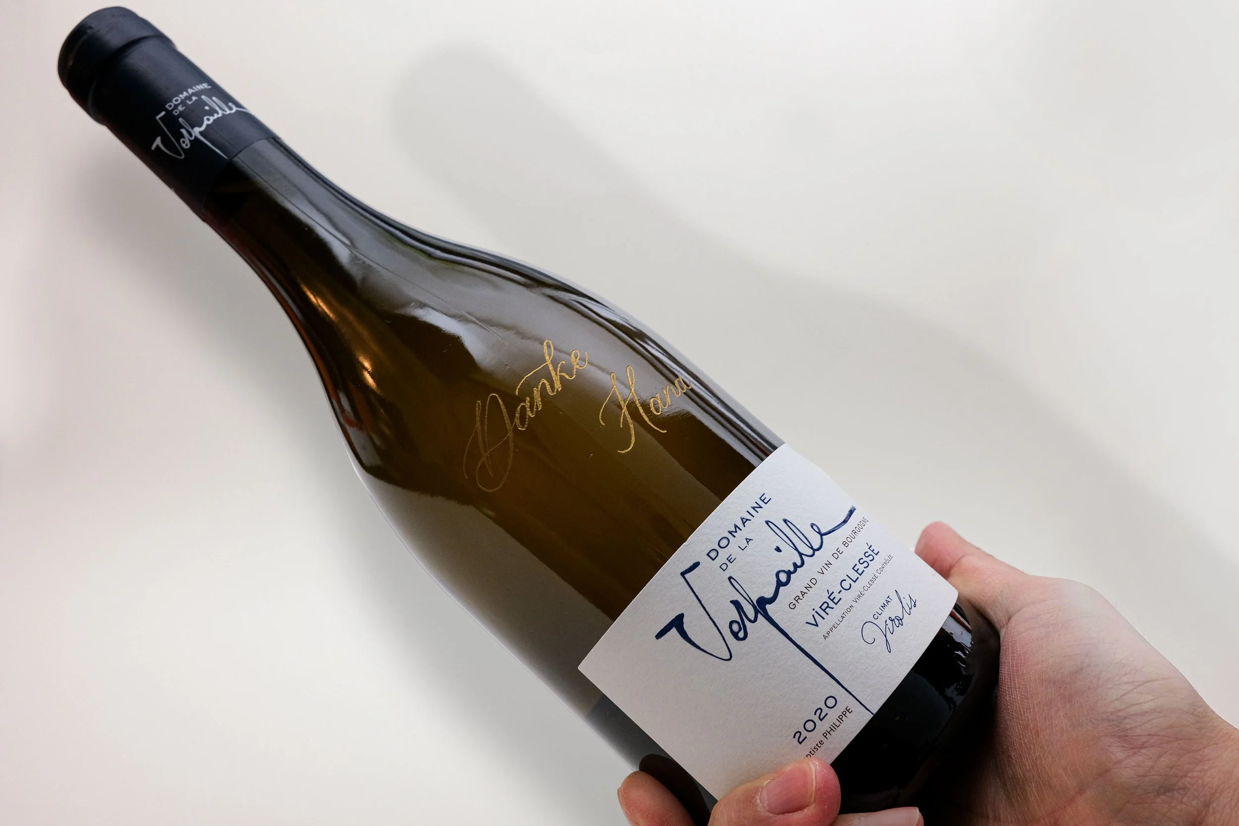Eine Weinflasche "Domaine de la Verpaille" mit einem weißen Etikett. Die Flasche wird in der Hand gehalten. Auf dem Etikett stehen Informationen wie "Viré-Clessé" und "2020". Man sieht auch eine handschriftliche Notiz auf der Flasche.