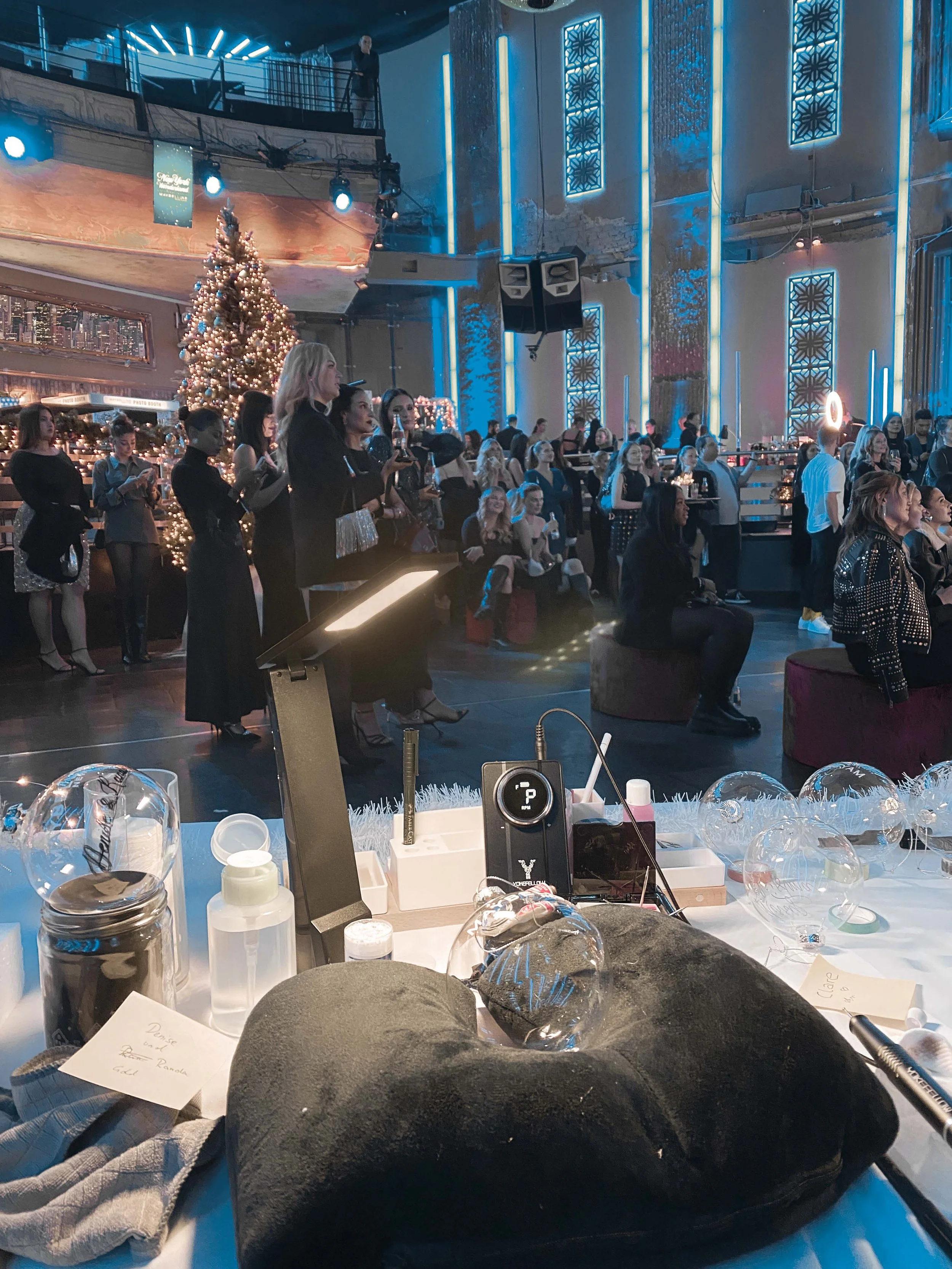 Innenansicht einer Veranstaltungshalle mit festlicher Dekoration, einem Weihnachtsbaum, vielen Menschen und einer Lichtshow. Im Vordergrund befindet sich ein Tisch mit verschiedenen Gegenständen wie Flaschen, Notizen und Dekorartikeln. Die Menschen scheinen aufmerksam zu sein, möglicherweise wird eine Präsentation oder Aufführung gezeigt.