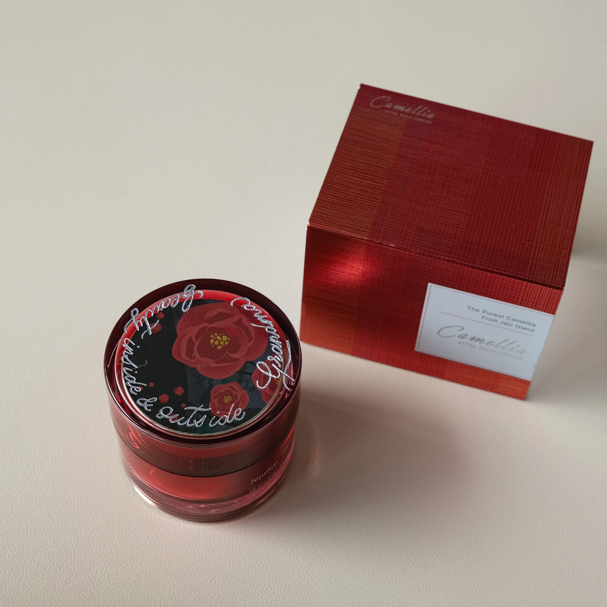Ein rotes Kosmetikprodukt mit einer Verpackung. Auf dem Deckel ist ein Blumenmuster zu sehen. Neben dem Produkt steht eine passende rote Schachtel mit der Aufschrift "Camellia Vital Rich Cream".