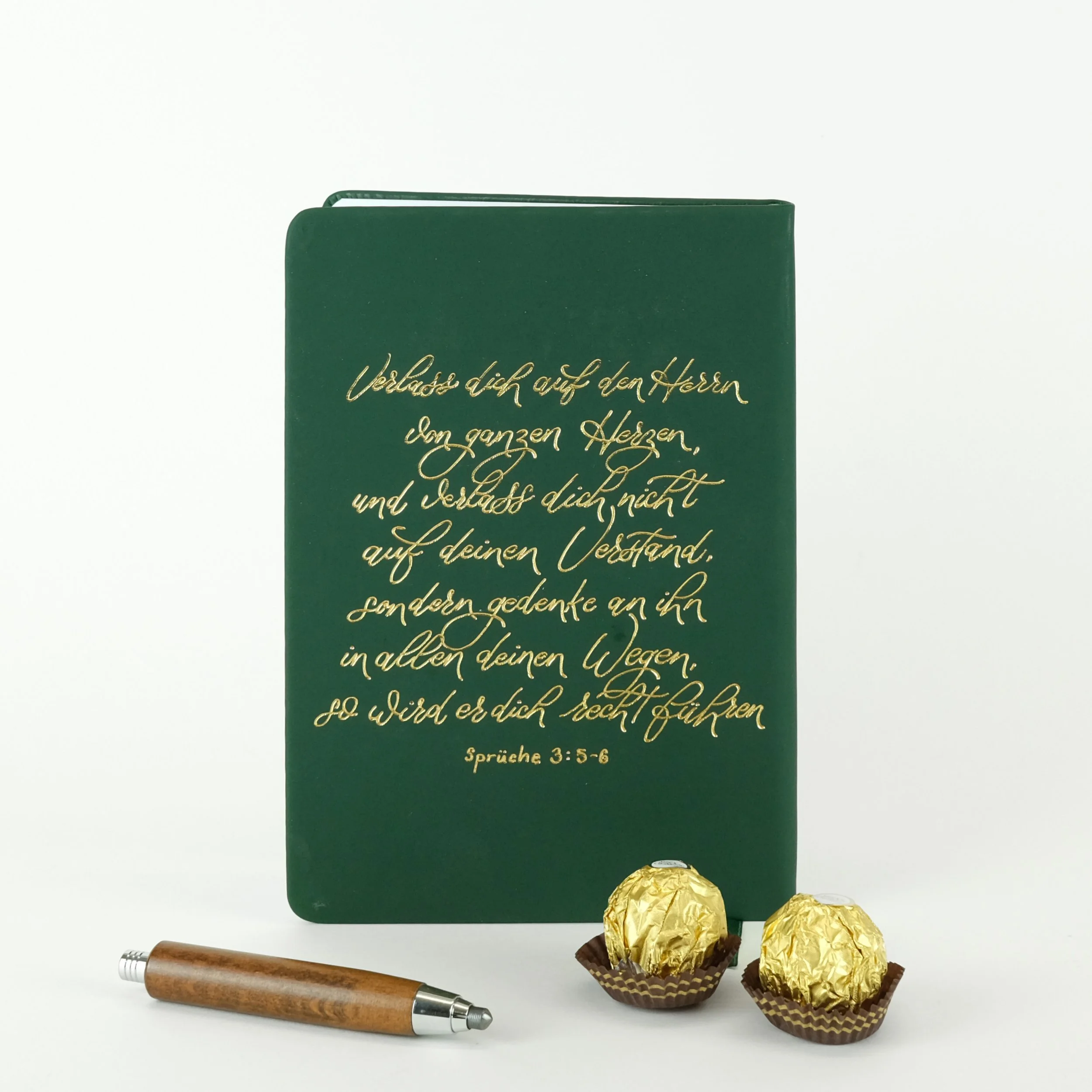 Grünes Notizbuch mit goldener Schrift und Bibelvers, daneben ein Kugelschreiber aus Holz und zwei Schokoladenpralinen in goldener Verpackung.