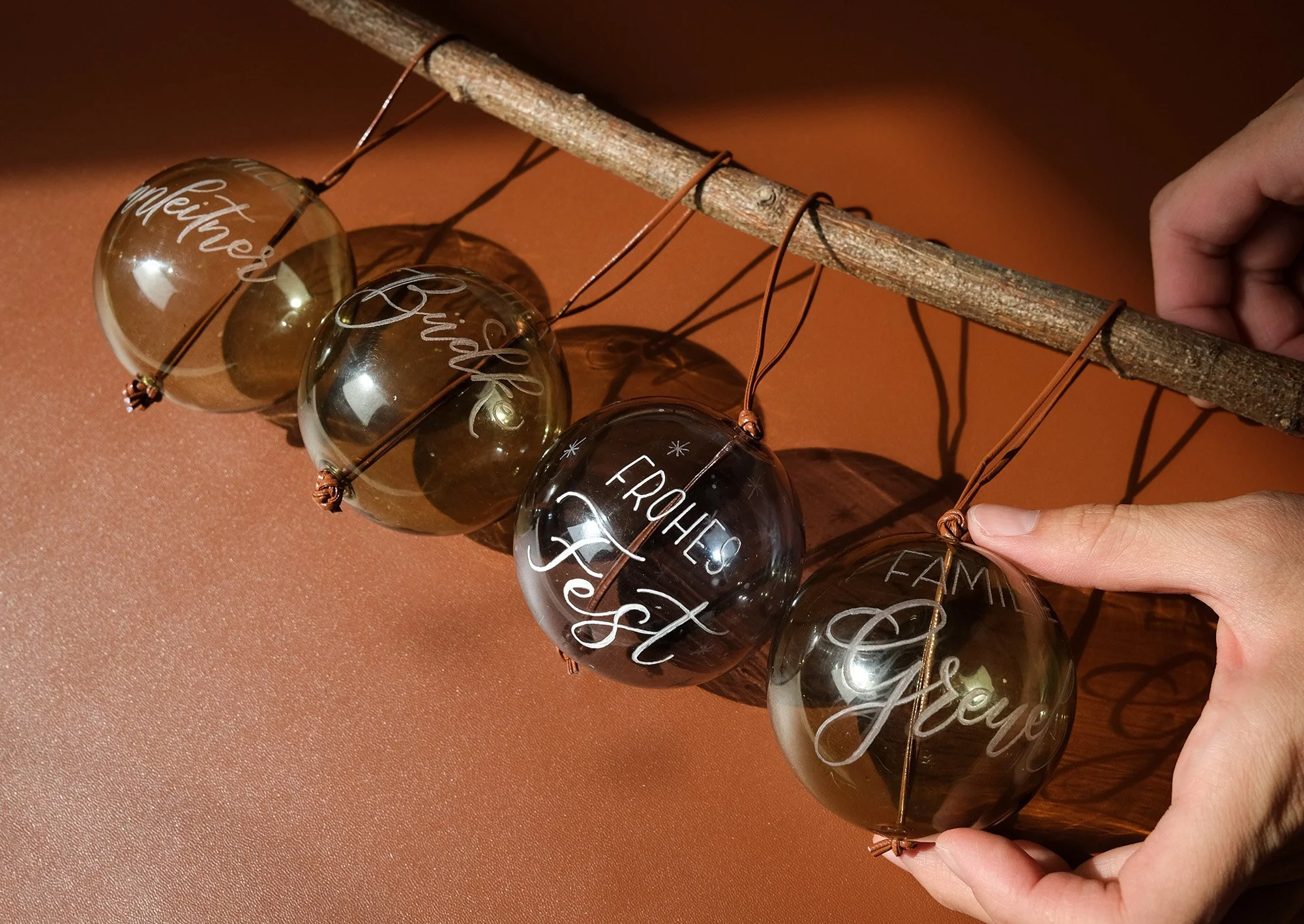 Vier transparente Weihnachtskugeln mit handgeschriebener Schrift, die an einem Ast hängen. Jede Kugel hat eine Inschrift, darunter "frohes Fest". Eine Hand hält die letzte Kugel. Hintergrund ist bronzefarben.