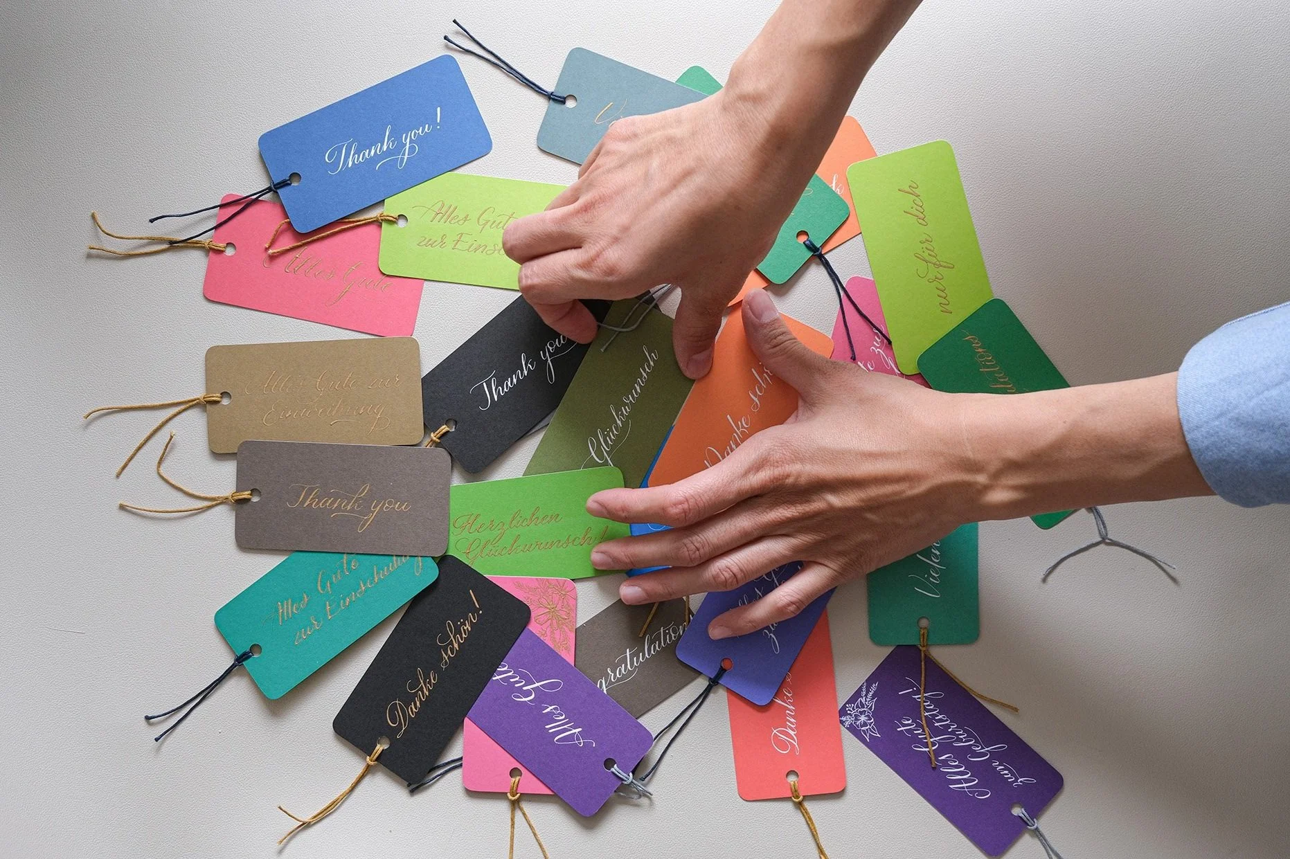Bunte Geschenkanhänger mit verschiedenen Aufschriften und Handschrift in unterschiedlichen Sprachen. Eine Hand sortiert die Anhänger auf einer hellen Oberfläche.