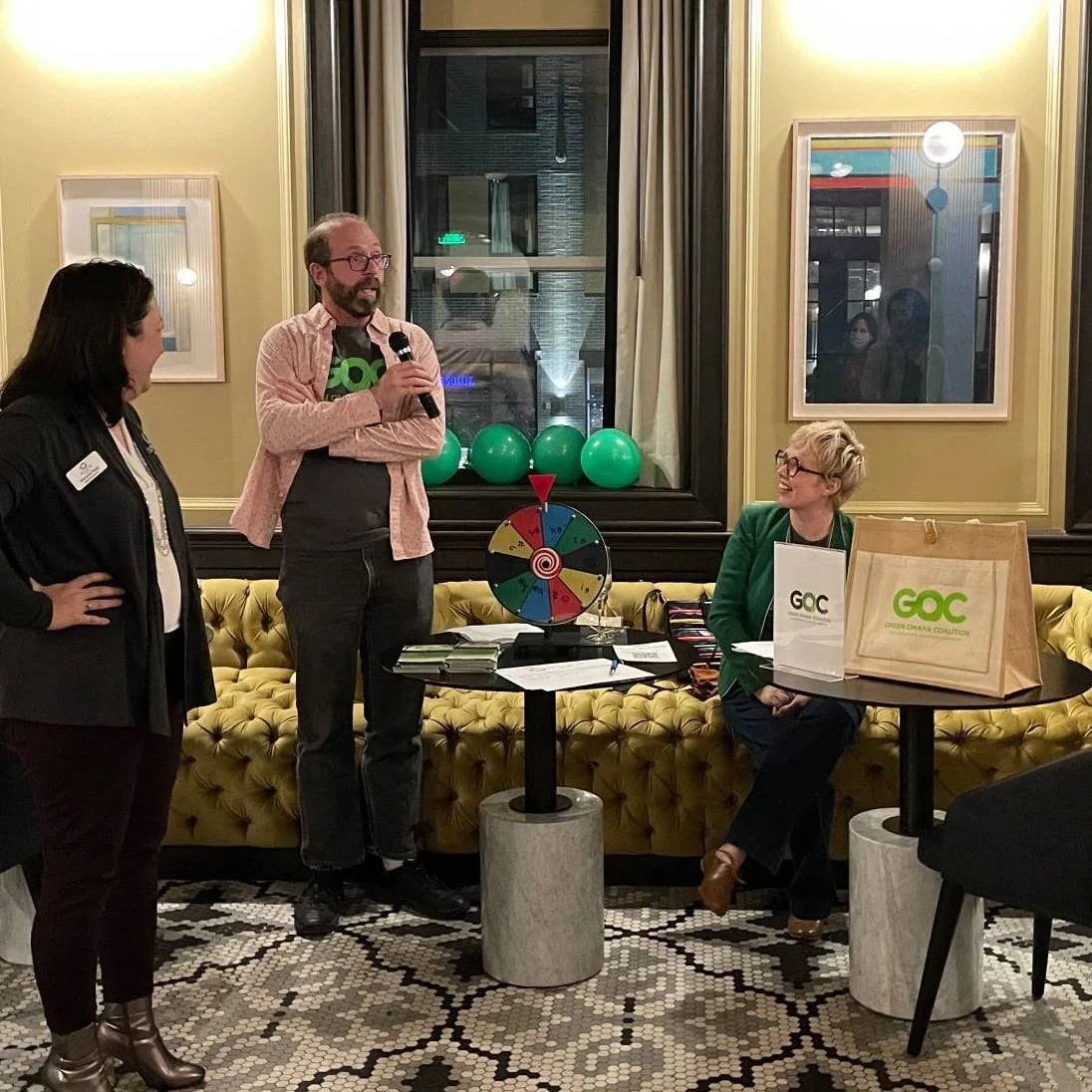 A man holding a microphone standing and speaking to two women in a room decorated with framed artwork, green balloons, and a colorful spinning wheel on a table. One woman is sitting at the table smiling, while the other woman is standing with her hands on her hips. There are GOC branded bags on the table.