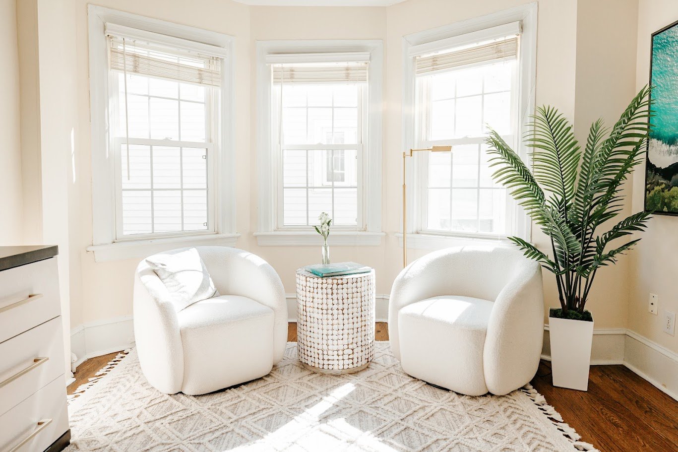 Light-filled sitting area with white accent chairs, textured rug, round side table, and faux palm tree.