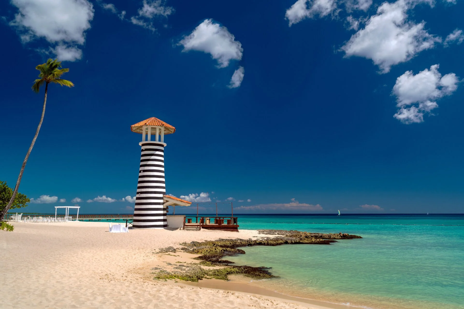 Tropical beach with a black and white striped lighthouse, a tall palm tree, turquoise ocean, sandy shore, and partly cloudy blue sky.