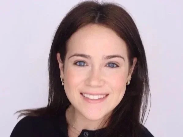 Portrait of a woman with dark brown hair, blue eyes, and a black top, smiling against a plain background.