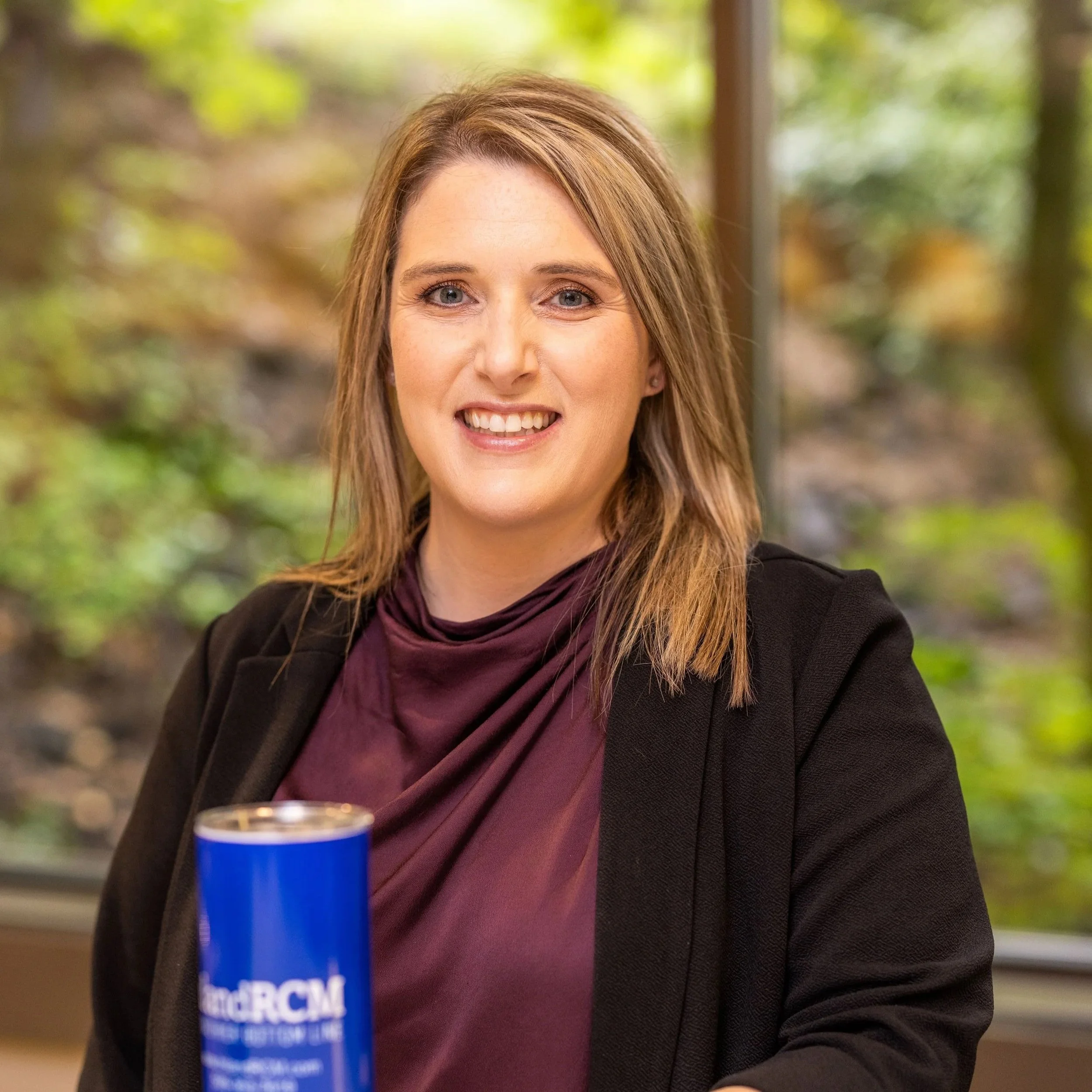 A woman with shoulder-length light brown hair, smiling at the camera, wearing a black blazer and a dark purple blouse, sitting indoors with a blurred green outdoor background visible through a window.