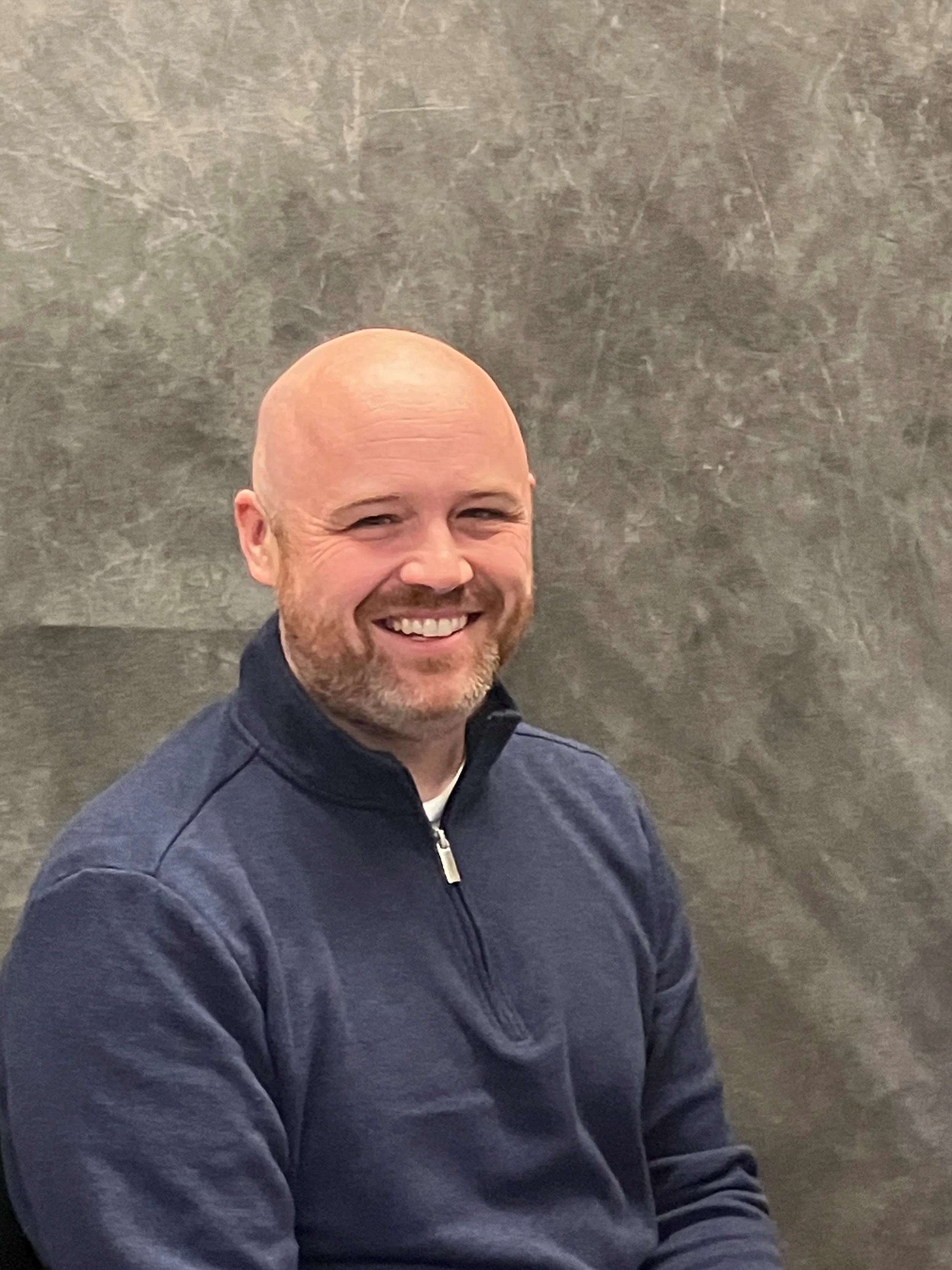 A smiling bald man with a beard, wearing a navy blue quarter-zip sweater, sitting against a gray textured wall.
