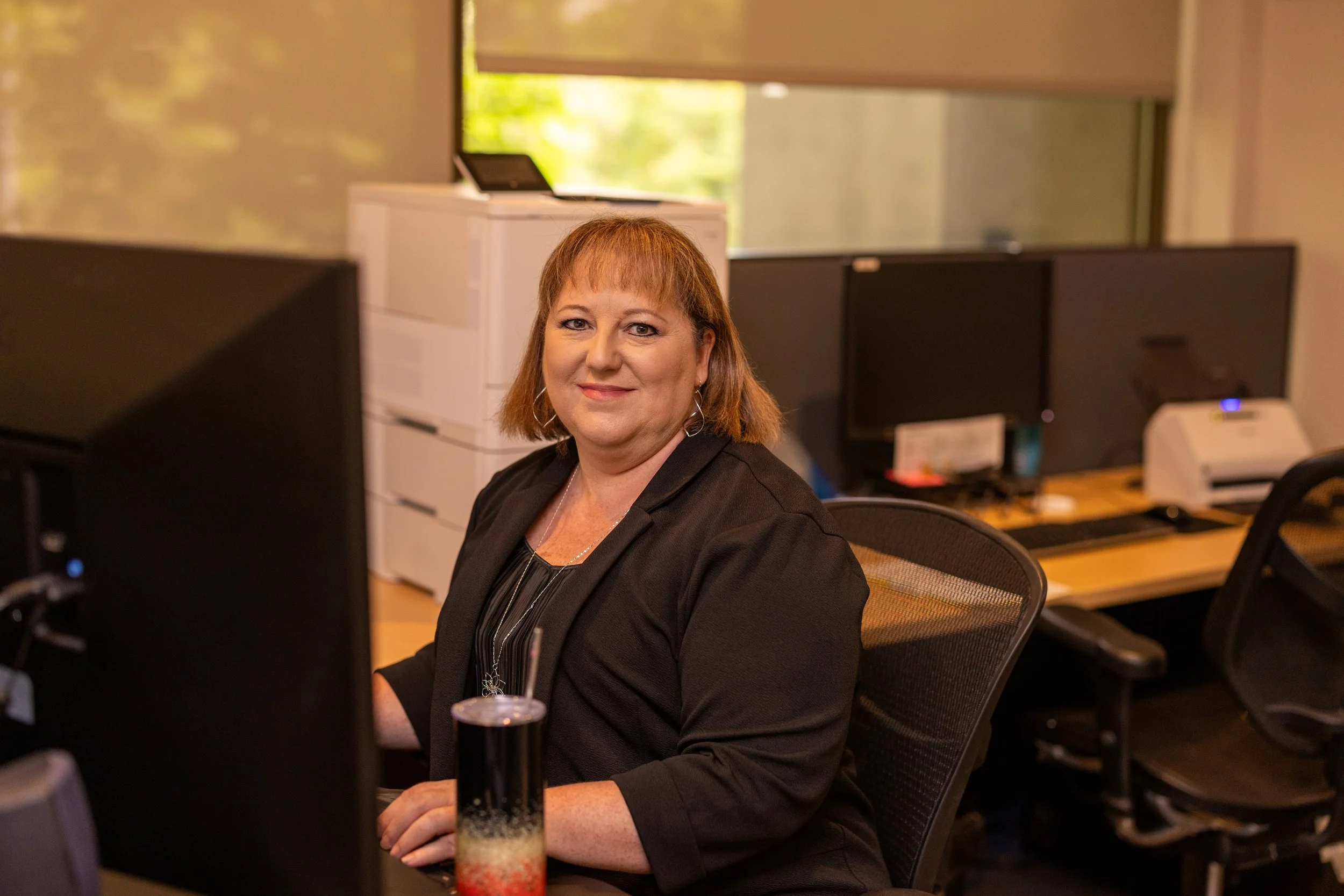 A woman with reddish-brown hair, dressed in a black blazer, sitting at a desk in an office with computer monitors, a printer, and office supplies.