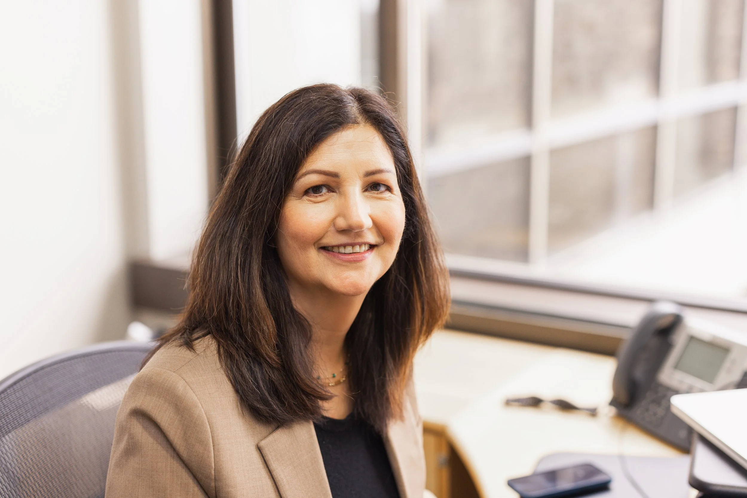 Smiling woman sitting at an office desk with a phone, computer, and large window in the background.