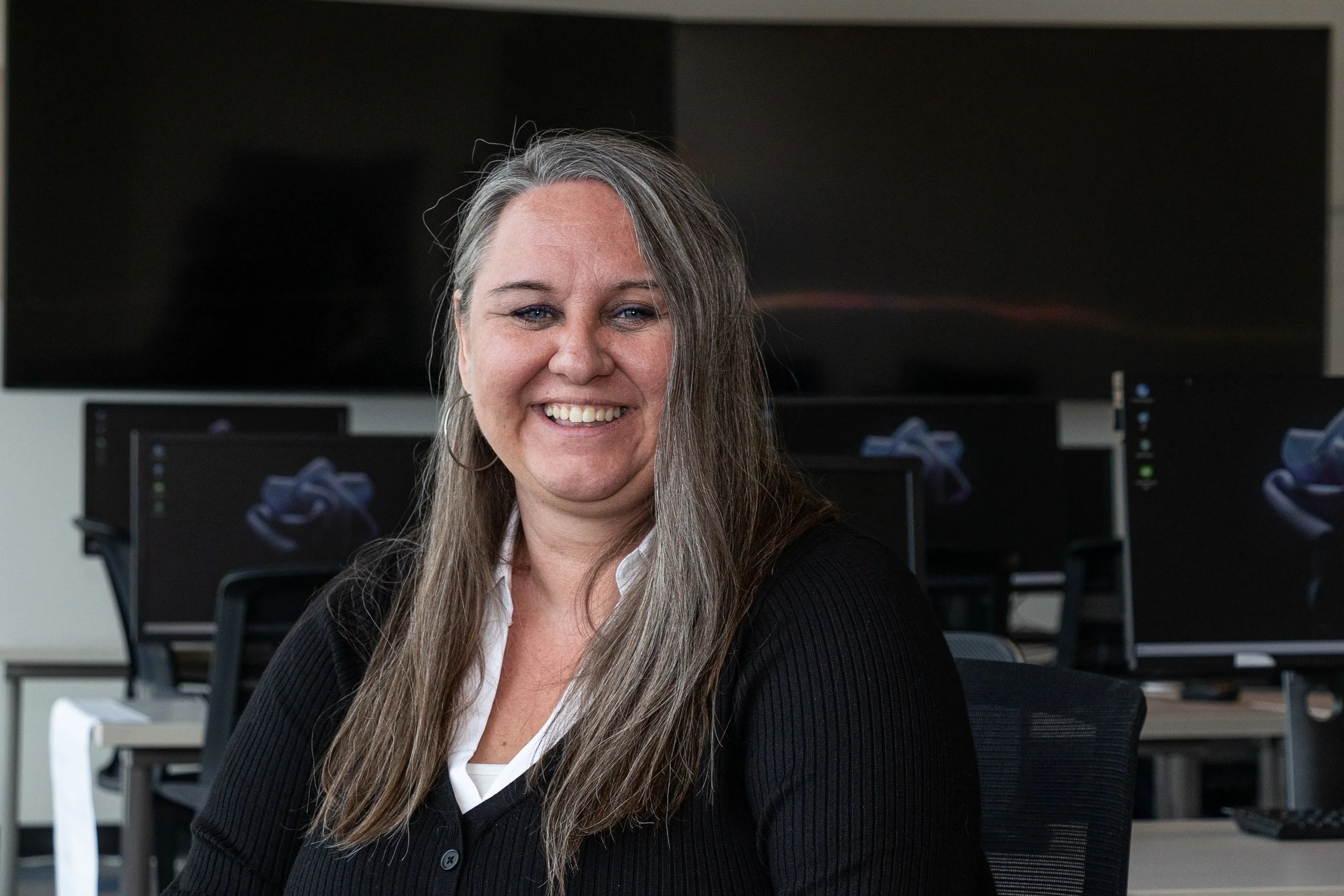 A smiling woman with long gray hair sitting in an office with multiple computer monitors behind her.