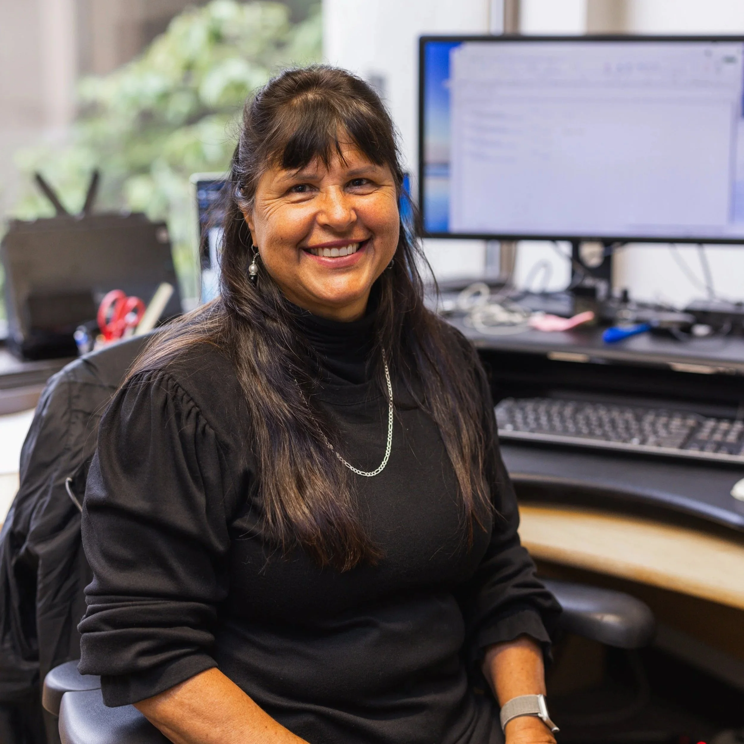 A woman sitting in an office, smiling at the camera, with dual computer monitors and office supplies on her desk.