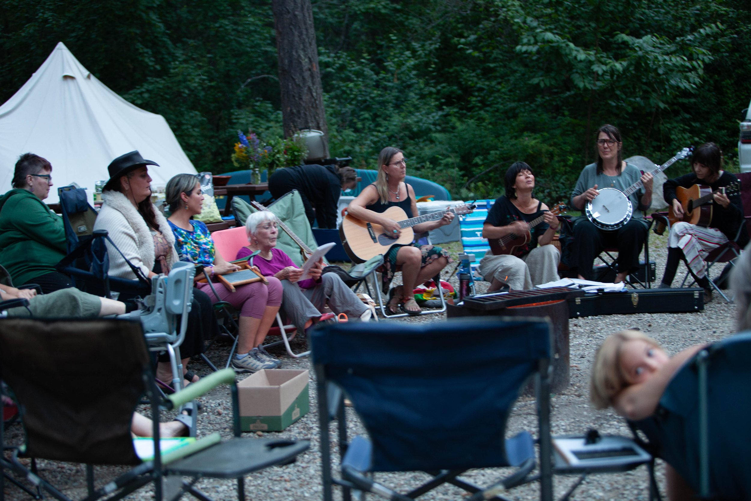Group of people sitting outdoors in a forested area, some playing guitars and singing, others listening, with tents and camping gear around.