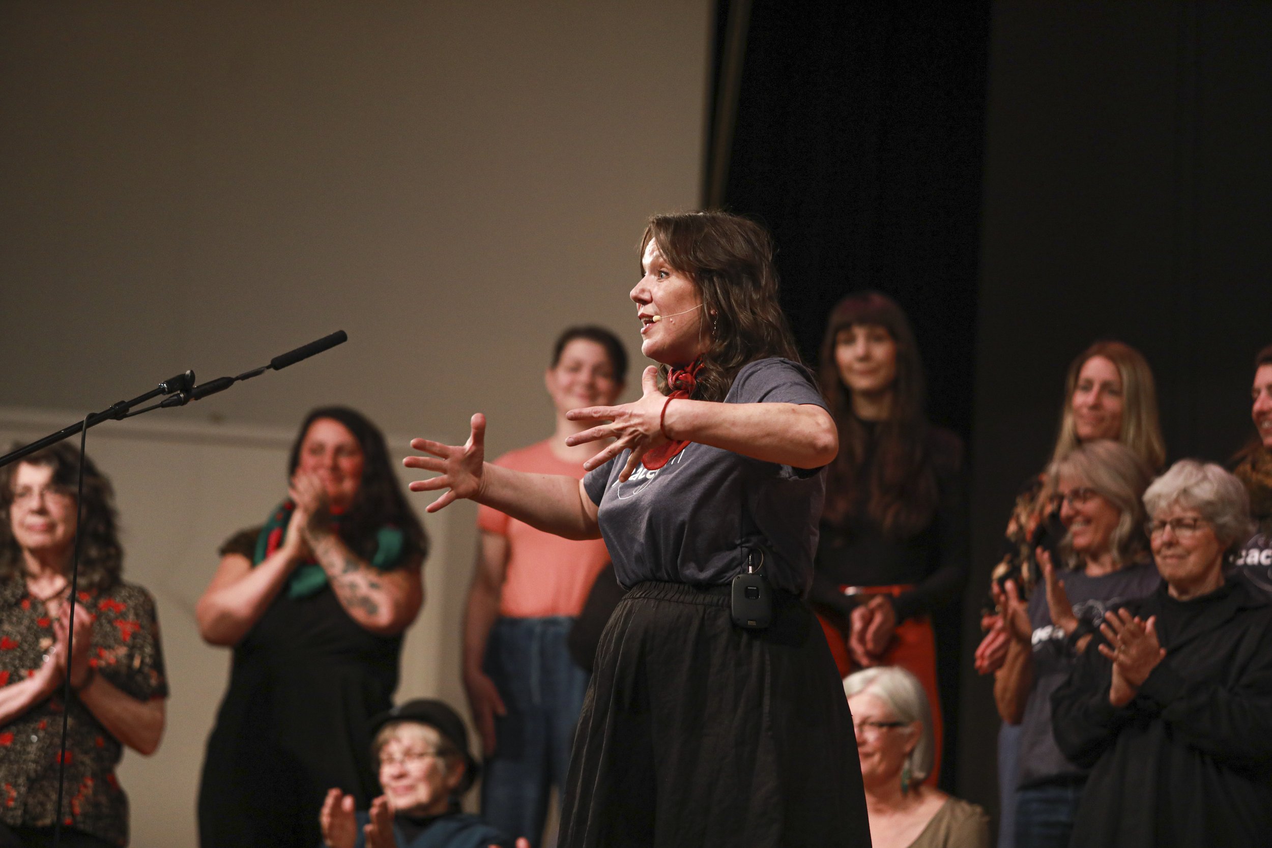 A woman speaking at a microphone on stage while a group of people behind her claps and smiles.