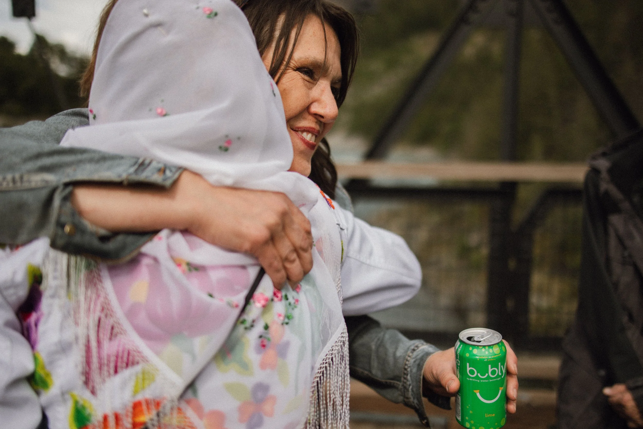 A woman and a person are embracing. The woman is smiling, and the person holding a green can of bubbly sparkling water is showing only their hand. The background is outdoors with trees and a fence.