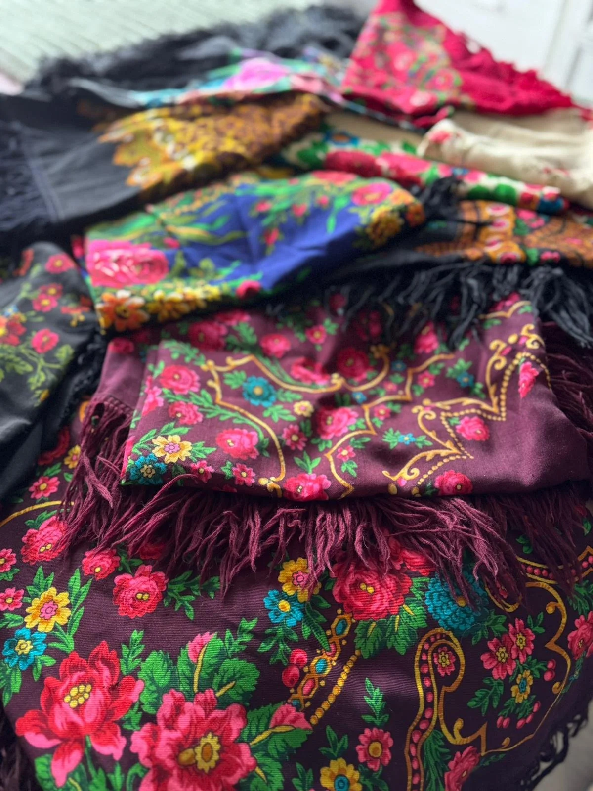 Assorted colorful scarves with floral patterns, textures, and fringes, arranged on a surface.