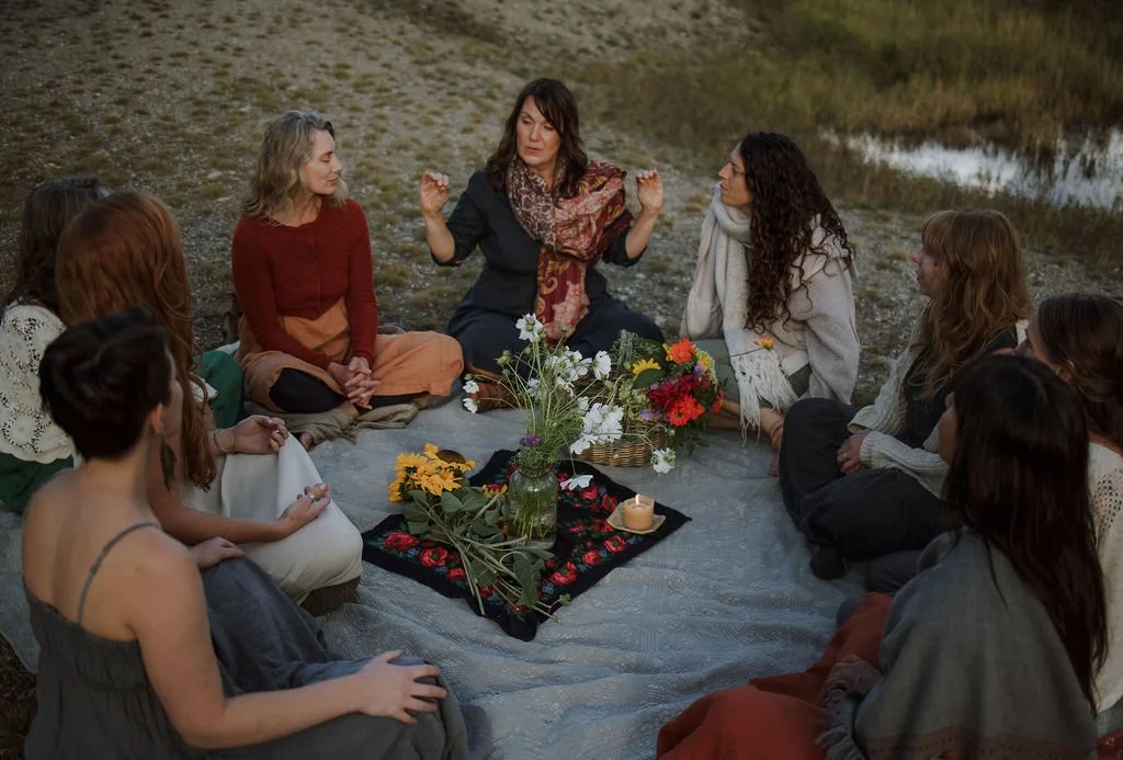 A group of women sitting cross-legged on blankets outdoors near a body of water, participating in a meditation or spiritual gathering with flowers and candles in the center.