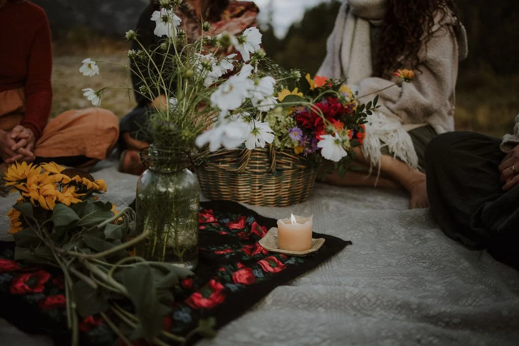 People sitting on a quilt outdoors with flower arrangements, a lit candle, and autumn clothing.