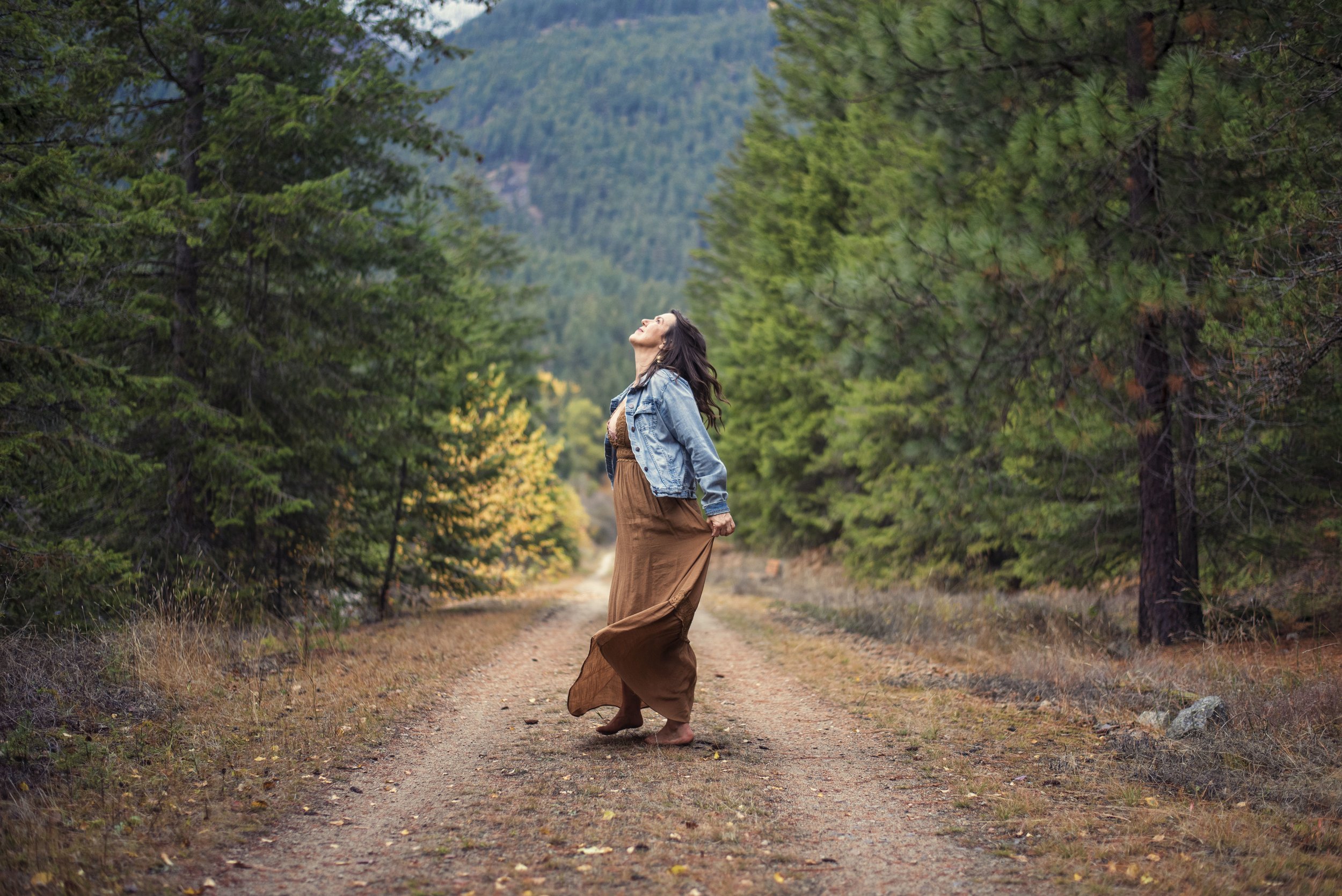 Woman with long dark hair in a denim jacket and brown flowing skirt standing on a dirt trail in a forested area with mountains in the background.
