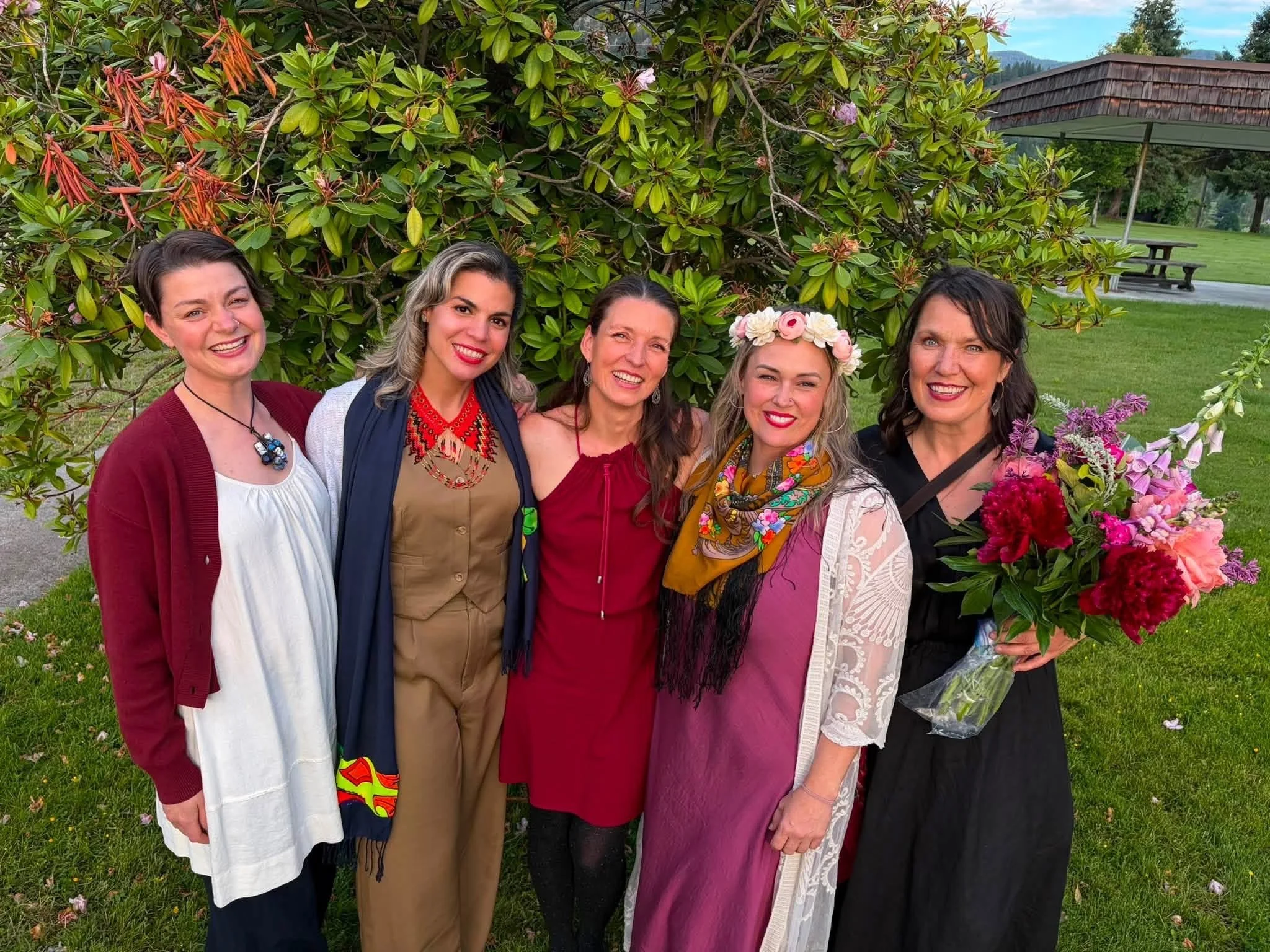 A group of six people, five women and one man, standing together outdoors in front of greenery, smiling. One woman is wearing a floral headband and holding a bouquet of flowers.