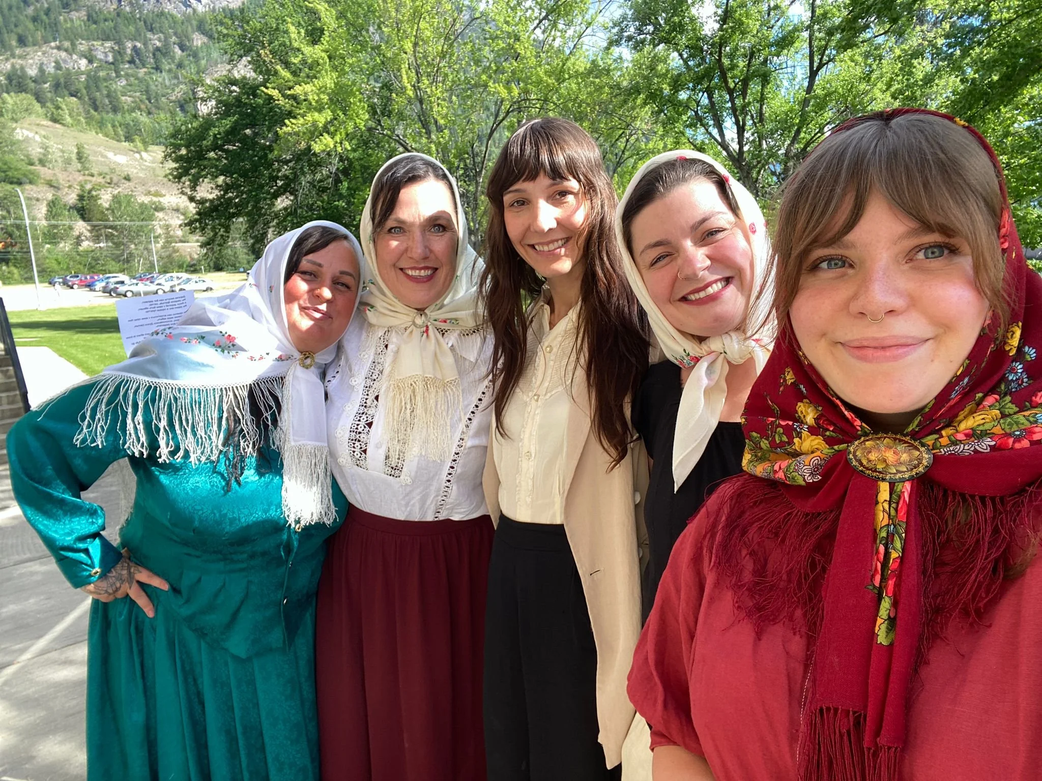 Group of five women outdoors wearing traditional clothing and headscarves, with a background of trees and blue sky.