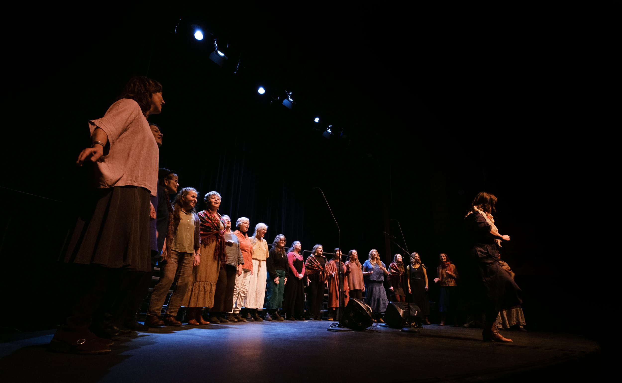A group of women standing on a stage, possibly during a choir or group performance, with a dark background and stage lights overhead.