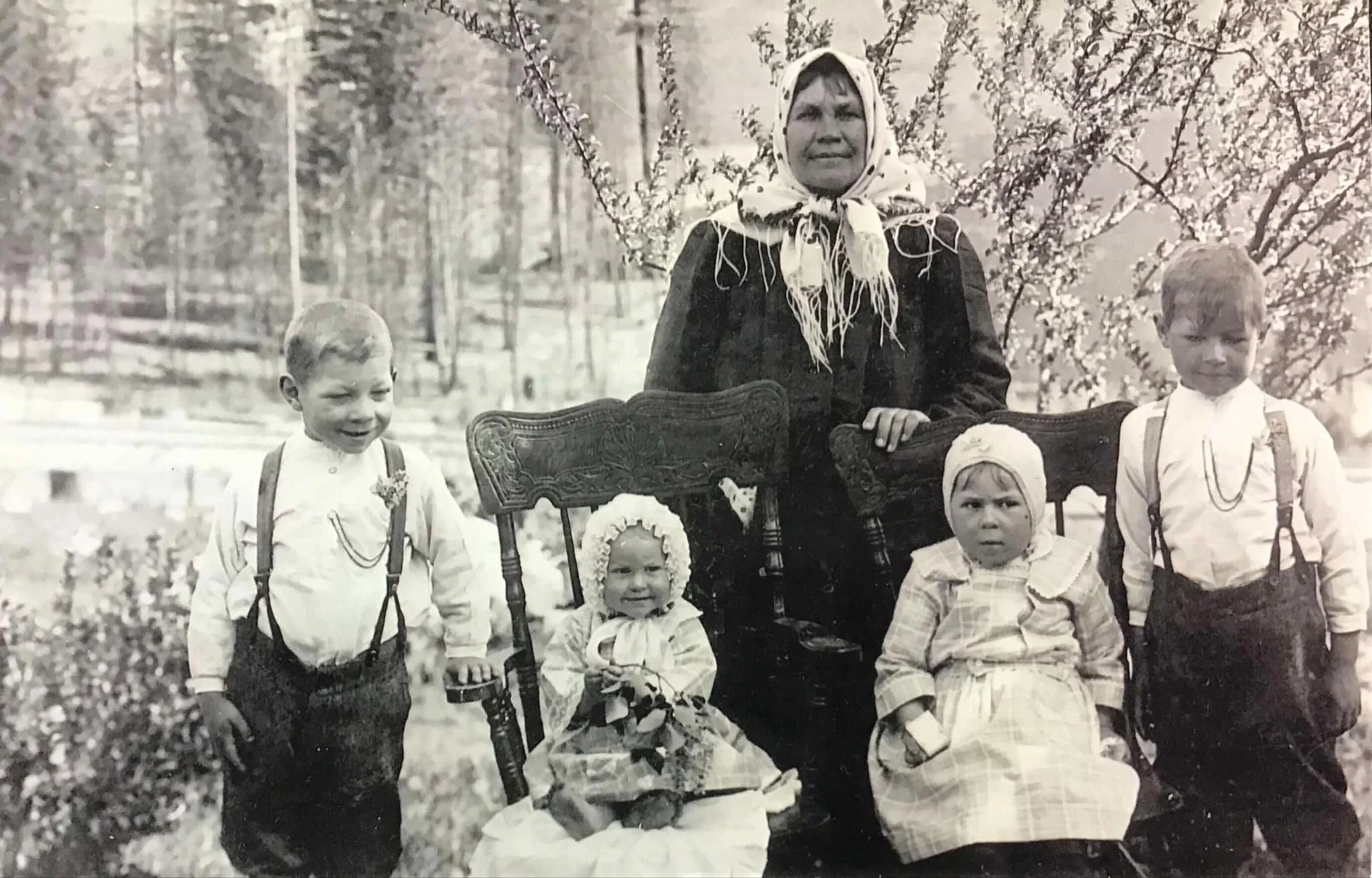 A black-and-white photo of a woman with children outdoors in a park or garden. The woman is standing behind a vintage wooden bench, wearing a headscarf and dark clothing. The children, dressed in old-fashioned clothing with suspenders and dresses, are gathered around her with some sitting and some standing. The background shows trees and floral bushes.